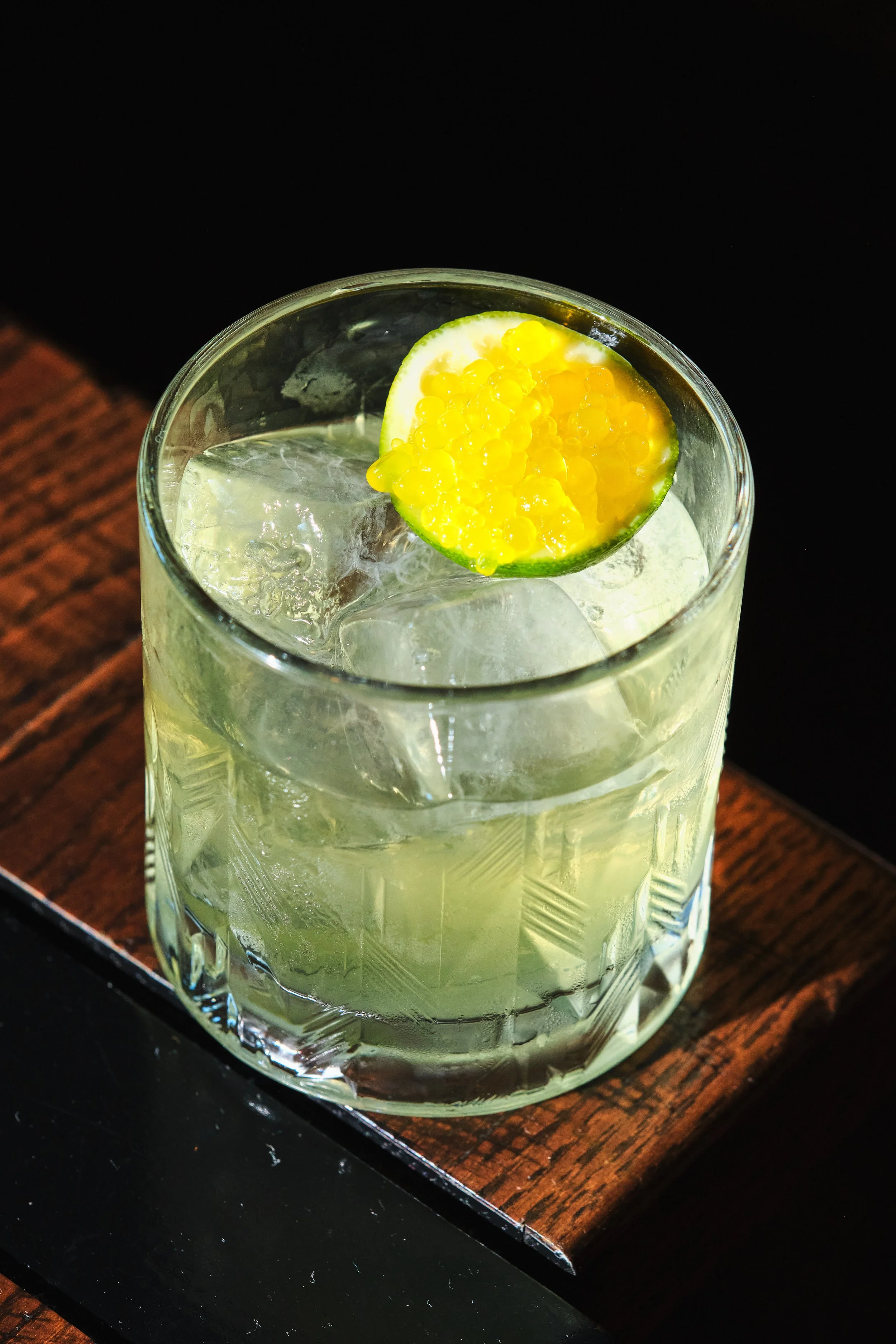 A clear glass of yellow-green cocktail with ice cubes, garnished with a slice of lime and yellow tapioca pearls, placed on a wooden coaster.
