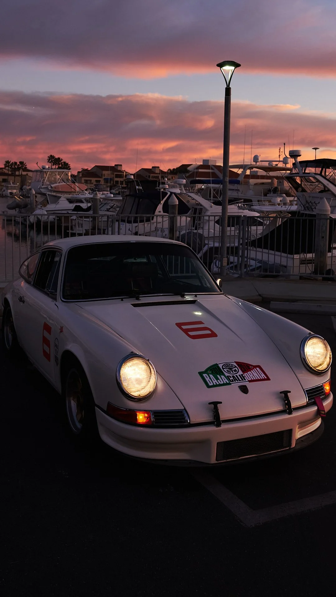 A classic white Porsche 911 race car with BMW M badge decals and number 6 on the side, parked near a marina during sunset with a sky of pink and purple clouds and boats docked in the background.