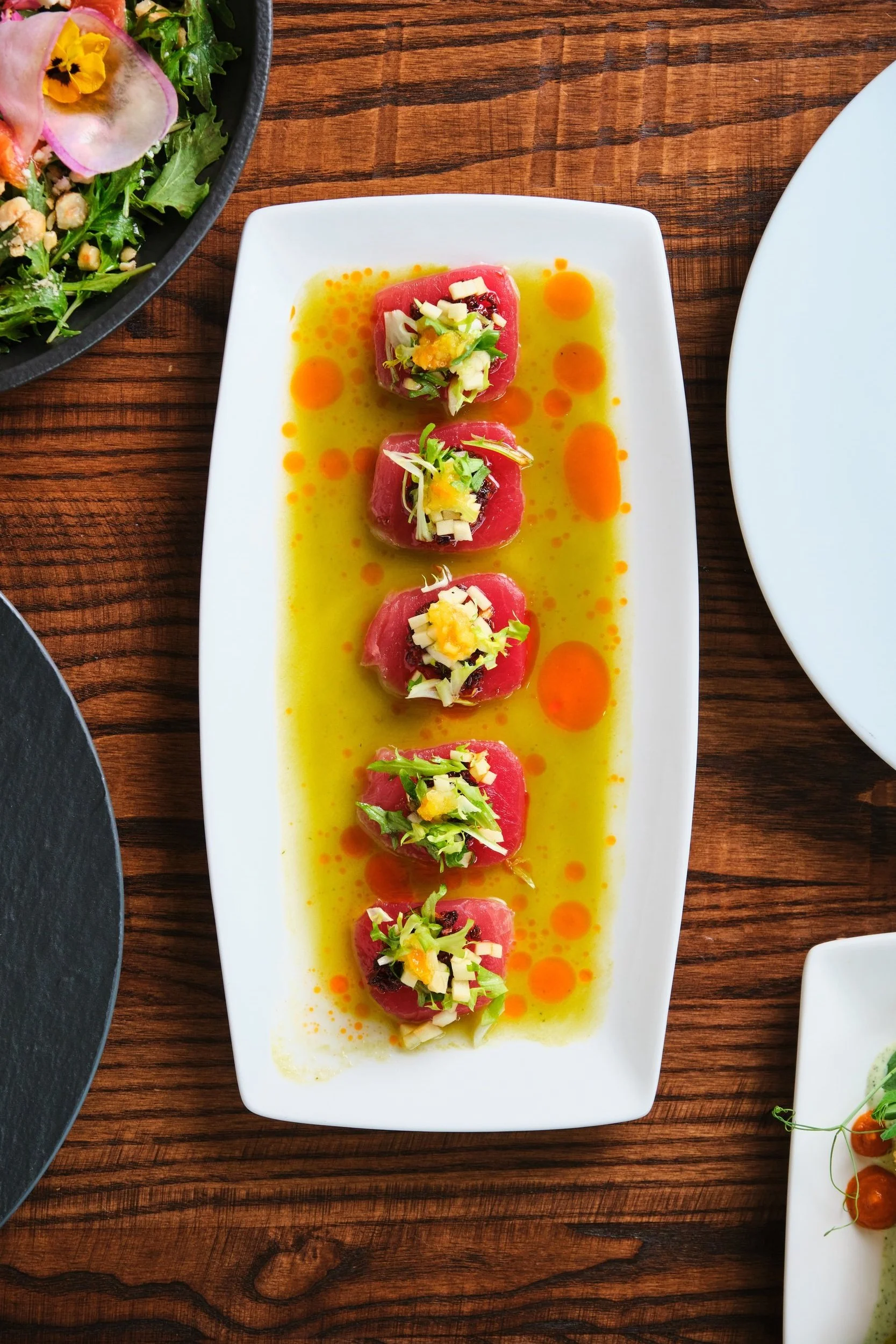 A rectangular white plate with five pieces of tuna tartare garnished with chopped vegetables and herbs, drizzled with yellow oil and orange sauce droplets, placed on a wooden table.