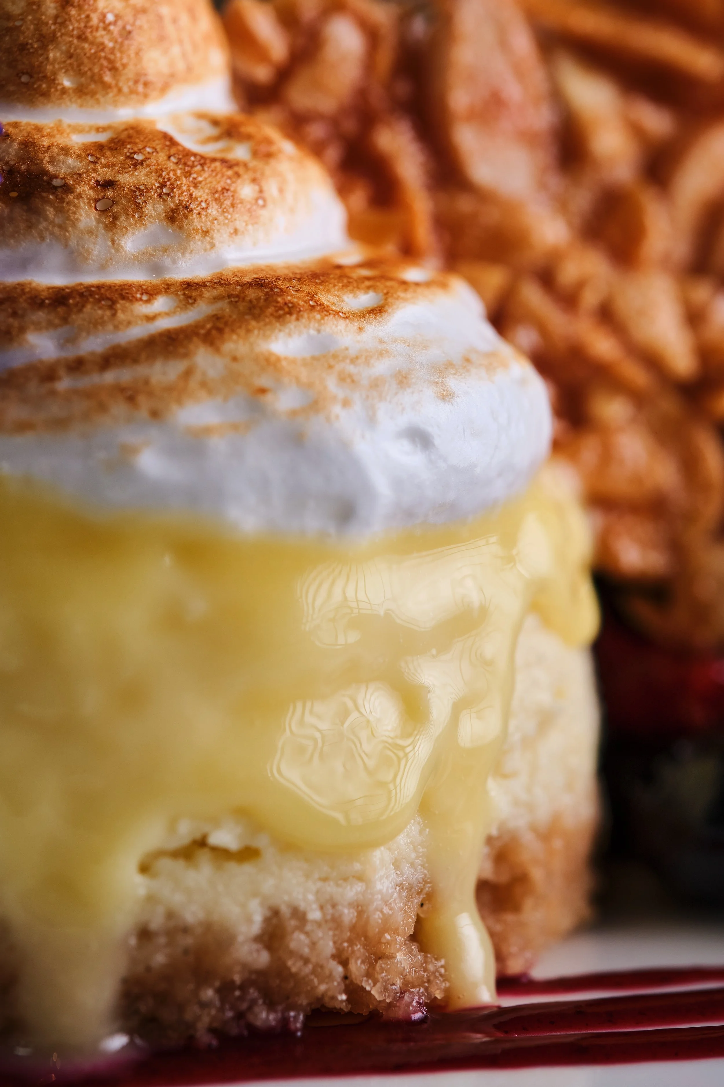 Close-up of a dessert with whipped cream, custard, and a baked topping, with caramelized nuts in the background.