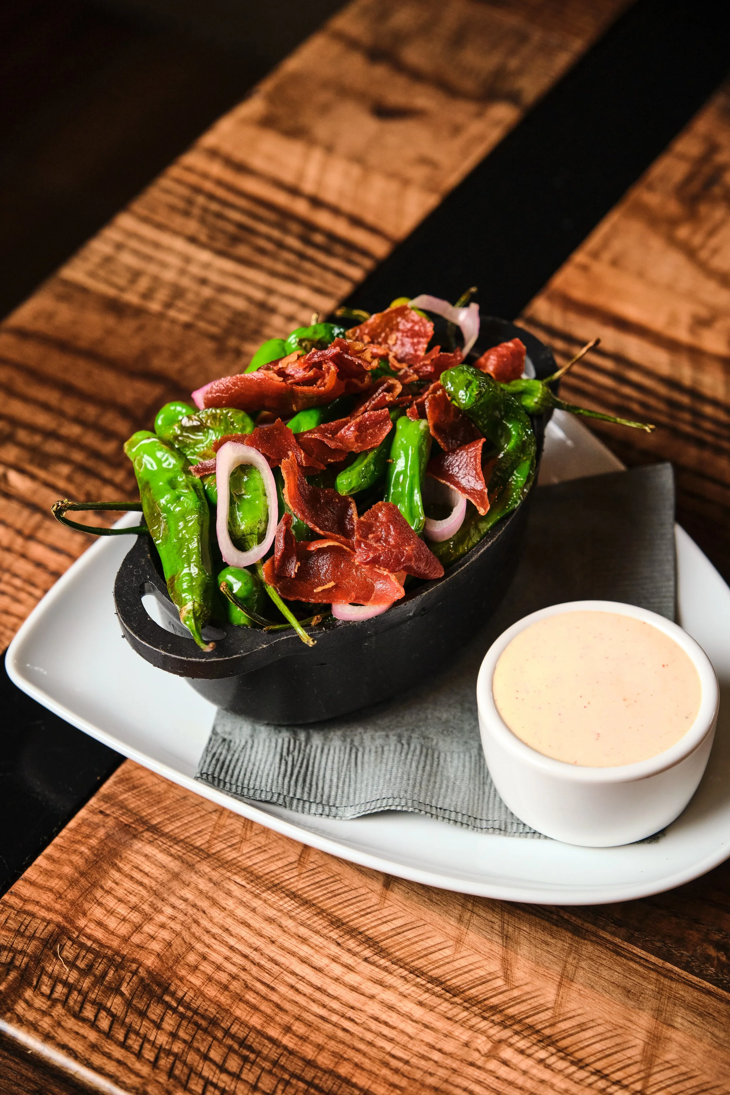 A dish of chiles, red onions, and shredded meat served in a black skillet accompanied by a small cup of creamy sauce on a white plate on a rustic wooden table.