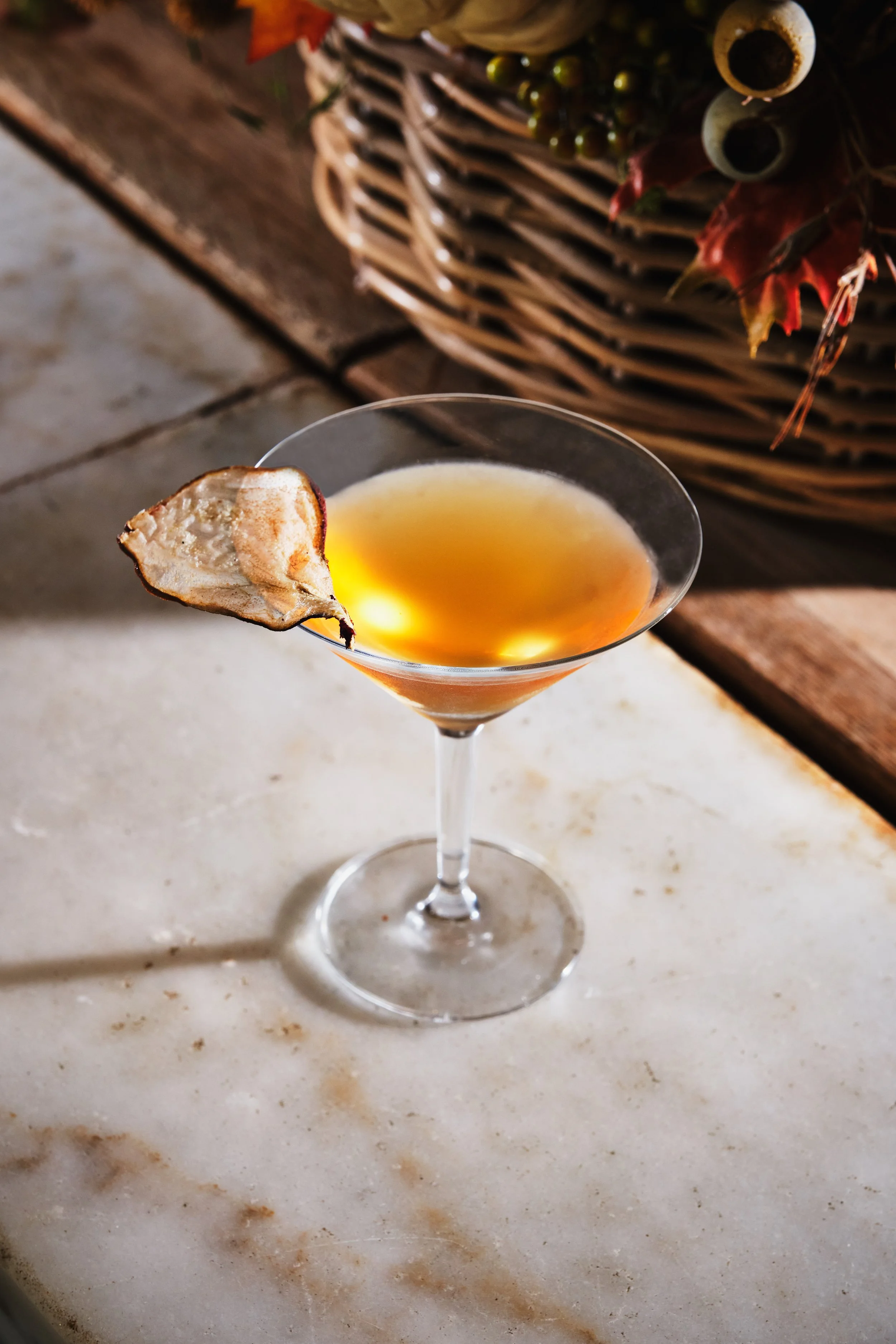 Cocktail in a martini glass with a dried pear slice garnish on the rim, sitting on a white marble surface with a wicker basket and autumn leaves in the background.
