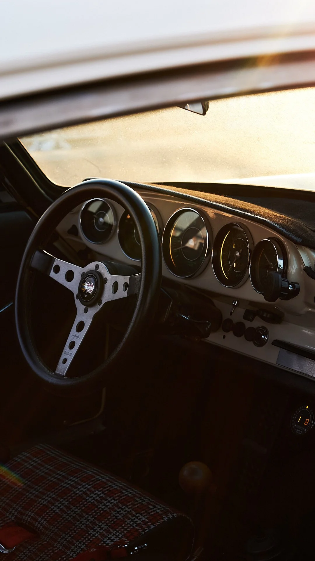 Inside a vintage car showing the steering wheel, dashboard, and plaid-patterned seat with sunlight coming through the windshield.