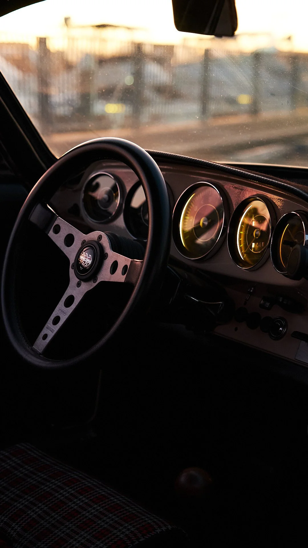 The interior view of a vintage car dashboard and steering wheel at sunset, with a blurred cityscape outside the windshield.