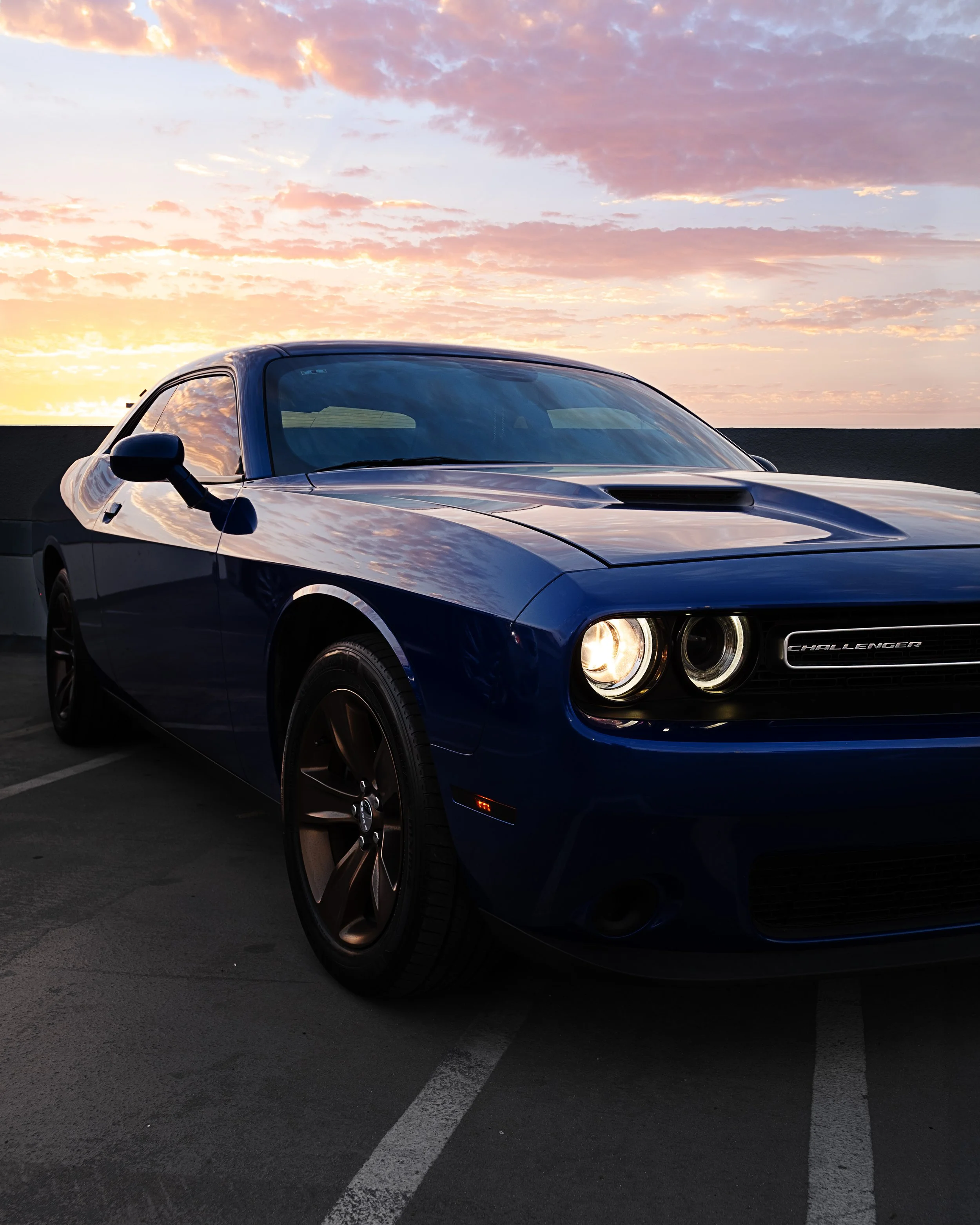 A blue Dodge Challenger parked on a rooftop parking lot during sunset with a sky filled with pink and orange clouds.