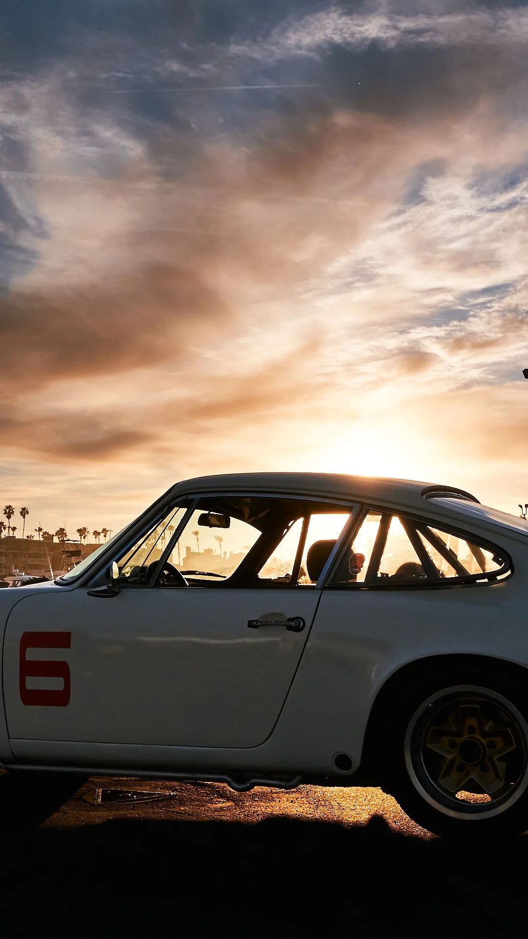 A white vintage racing car with the number 6 on its door parked at sunset with a sky filled with clouds and palm trees in the background.