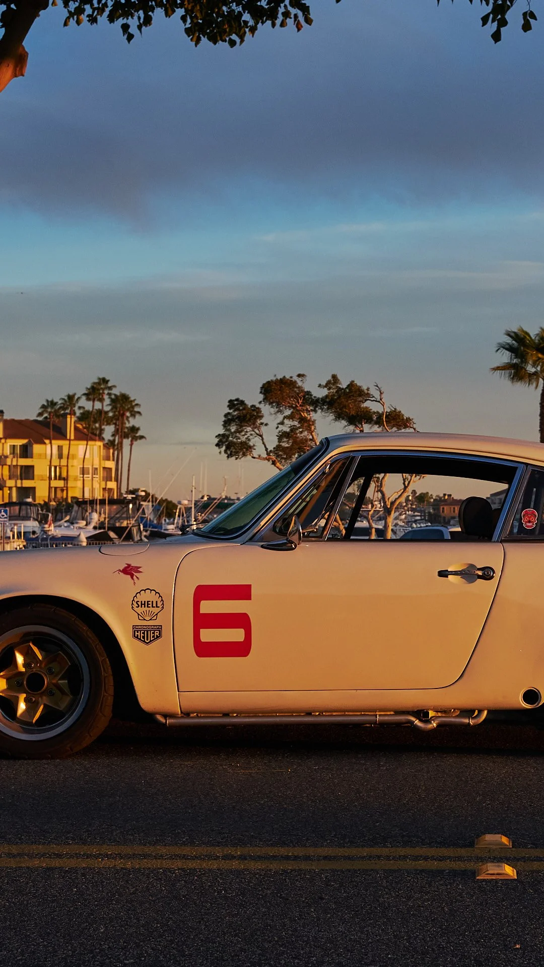 A vintage white race car with the number 6 painted in red on its door, parked on a road near a marina with boats and palm trees under a partly cloudy sky at sunset.