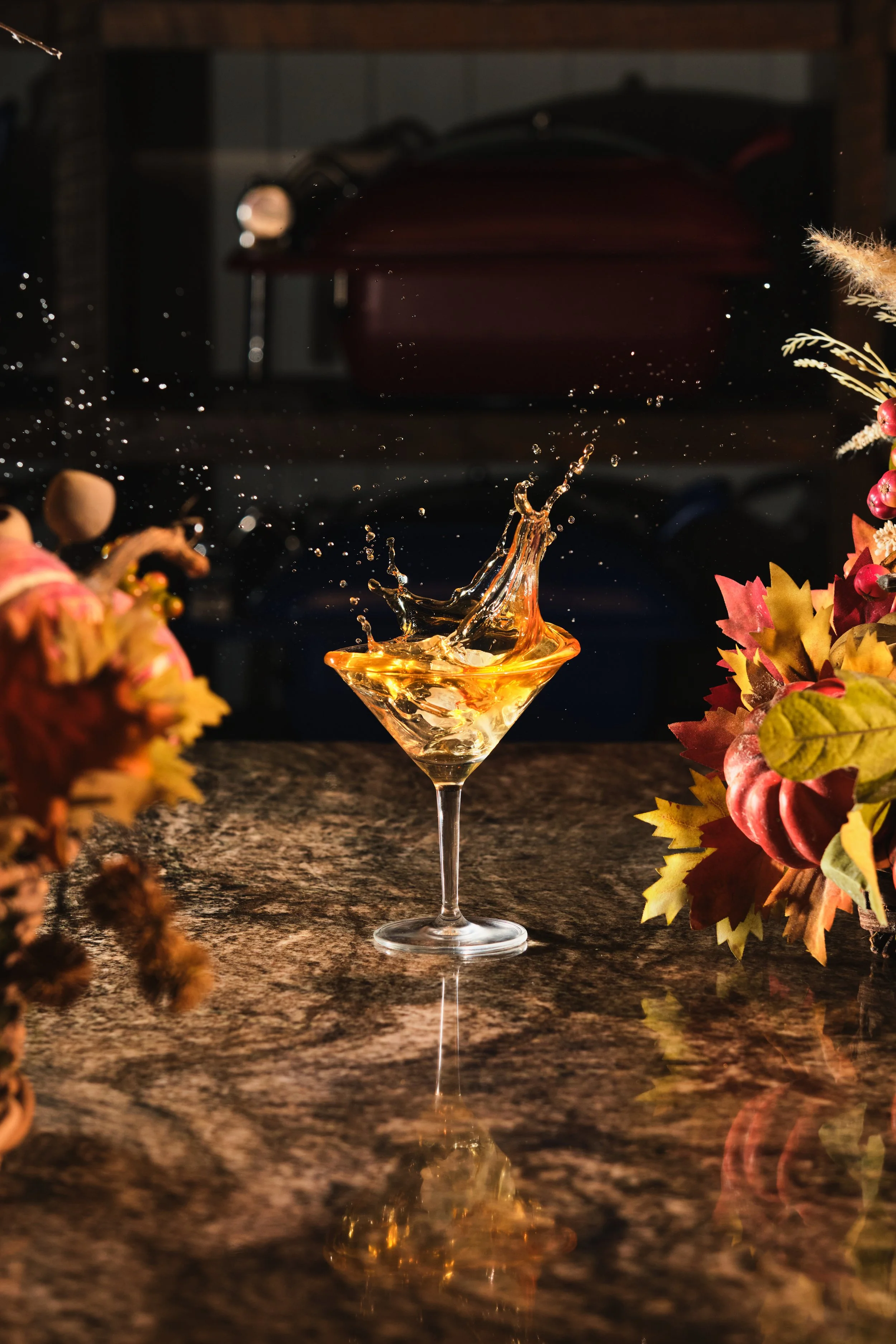 A splash of golden-colored liquid in a martini glass on a marble countertop, surrounded by autumn-themed foliage and decorations.
