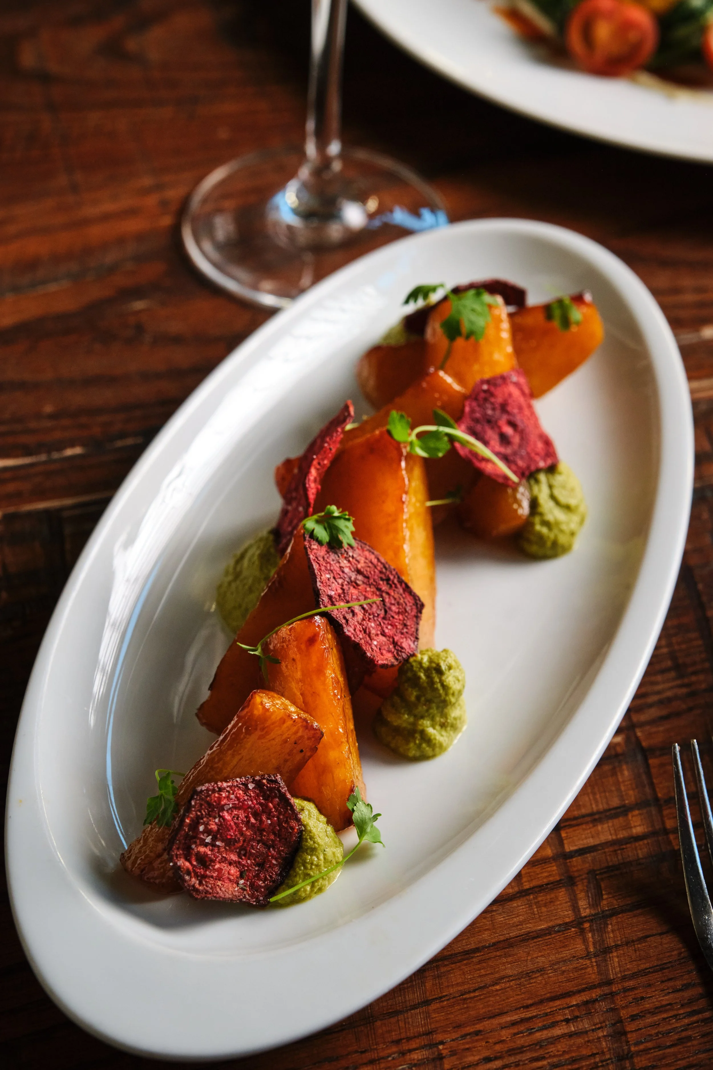 A white oval plate with roasted sweet potato pieces, red beet chips, dollops of green sauce, and garnished with microgreens, set on a wooden table with a wine glass and another dish in the background.