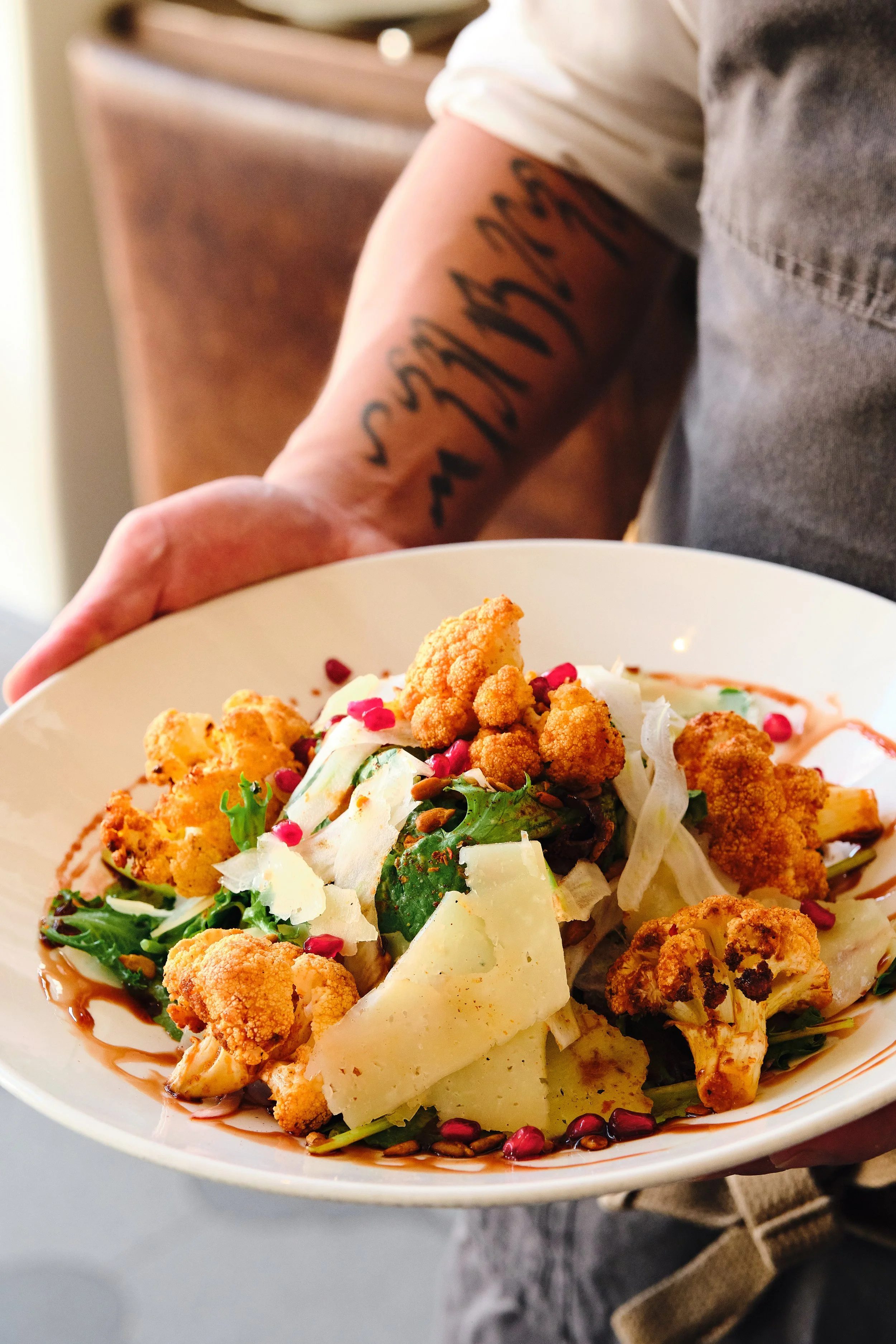 Person holding a white bowl of salad with cauliflower, cheese, mixed greens, pomegranate seeds, and dressing.
