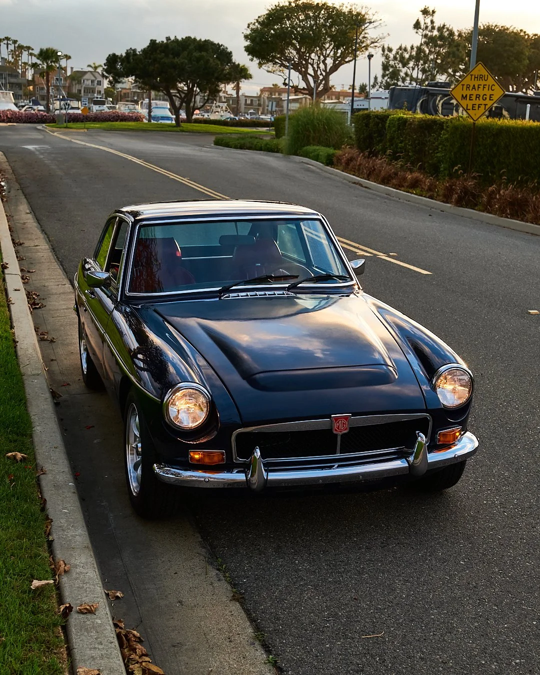 A classic black MG car parked on the side of a street, with green grass and trees, and marina with boats in the background during sunset.