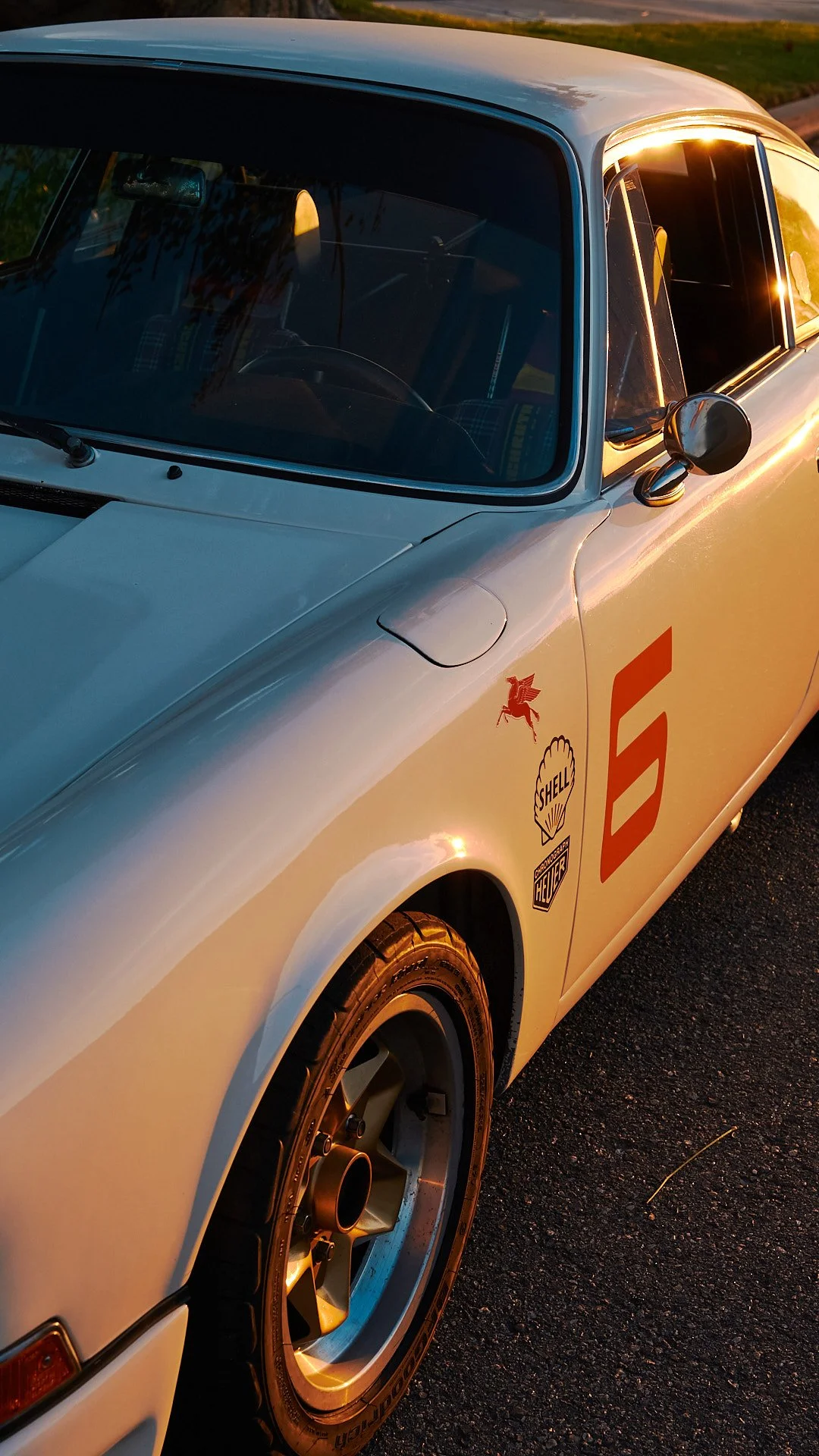 Close-up of a vintage race car with a white body, racing decals including the number 6, and sponsorship stickers like Shell and Heuer, parked on a dark asphalt surface during sunset.