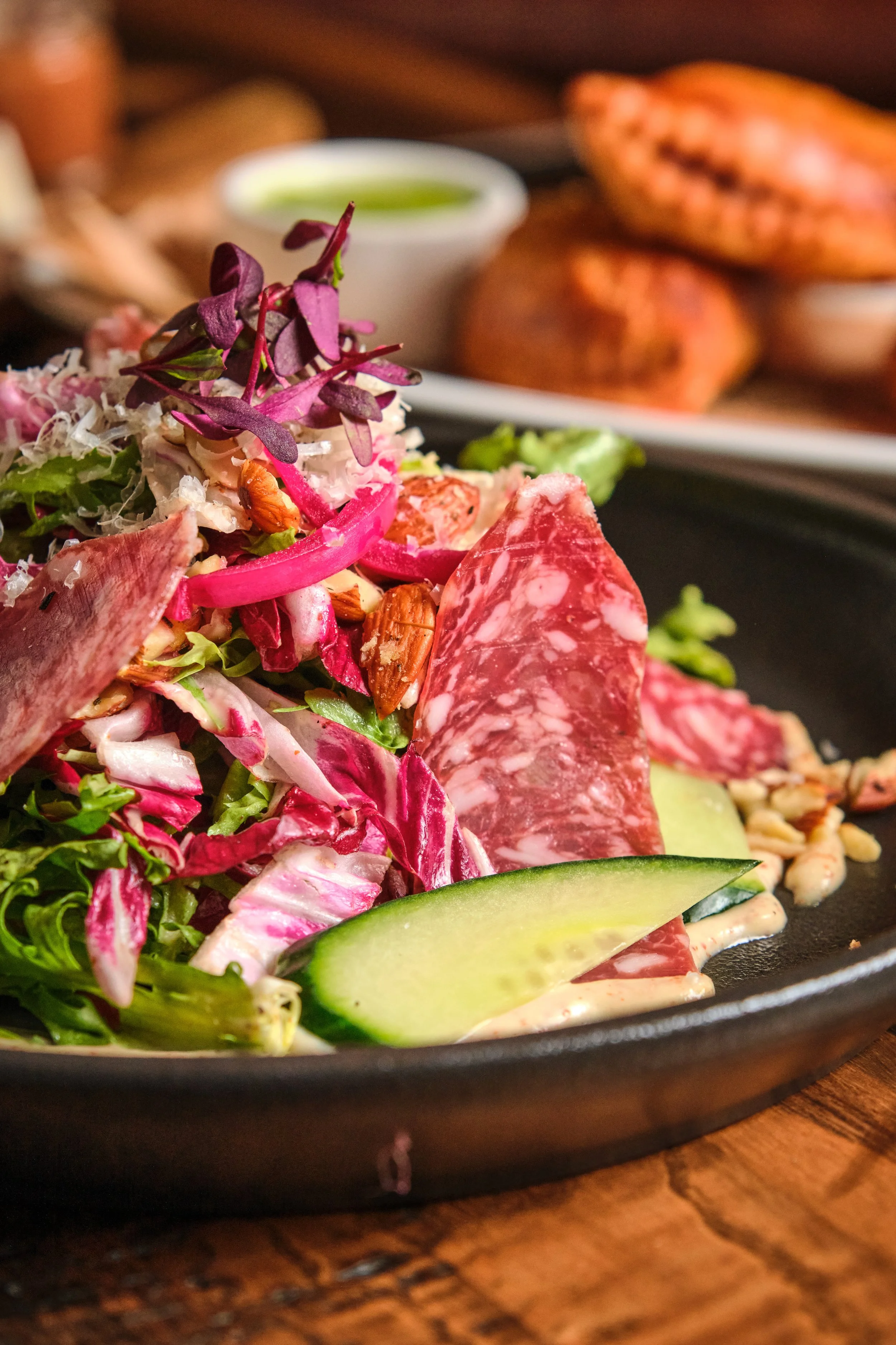 Close-up of a colorful salad with cured meat, cucumber, and purple microgreens on a black plate, with a side dish and dipping sauce in the background.