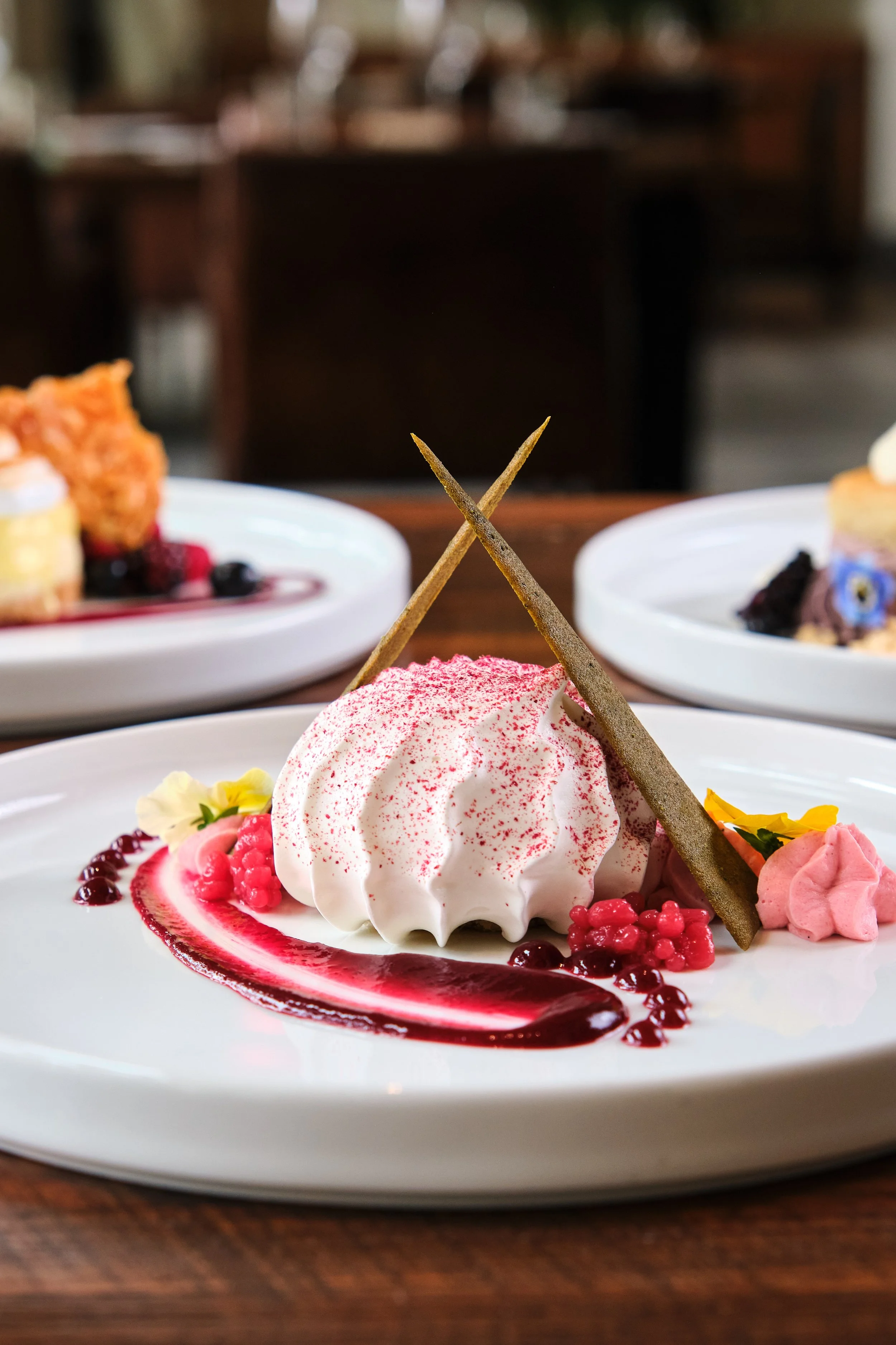 Elegant dessert plate with pink whipped cream, red berry sauce, decorative berries, yellow flowers, and two crossing brown sticks.