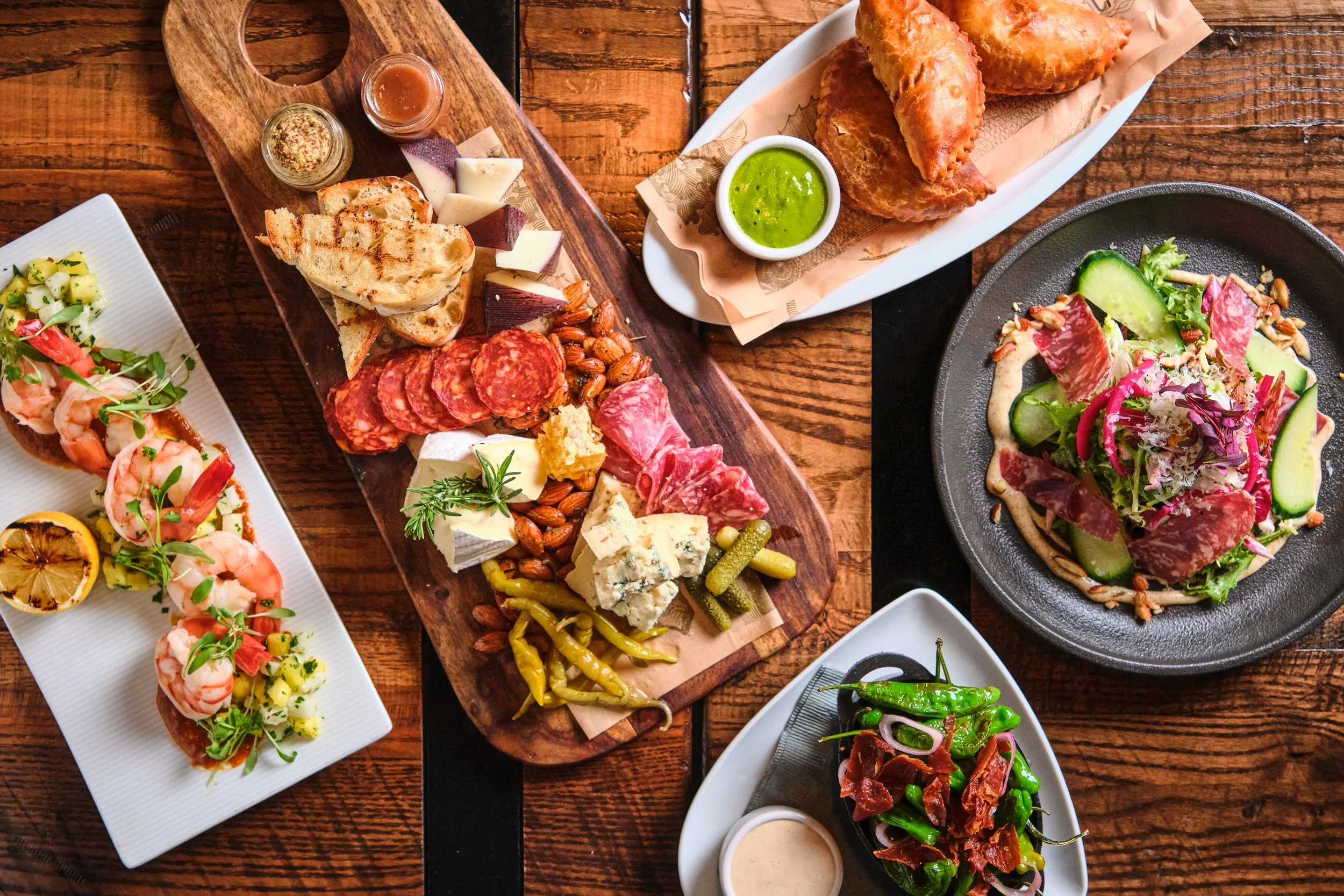 Assorted dishes including a charcuterie board with cheeses, meats, nuts, and bread, a plate of fried empanadas with green sauce, a Beet salad with cucumbers and greens, a shrimp canapé with lemon, and a roasted pepper and prosciutto salad on a rustic