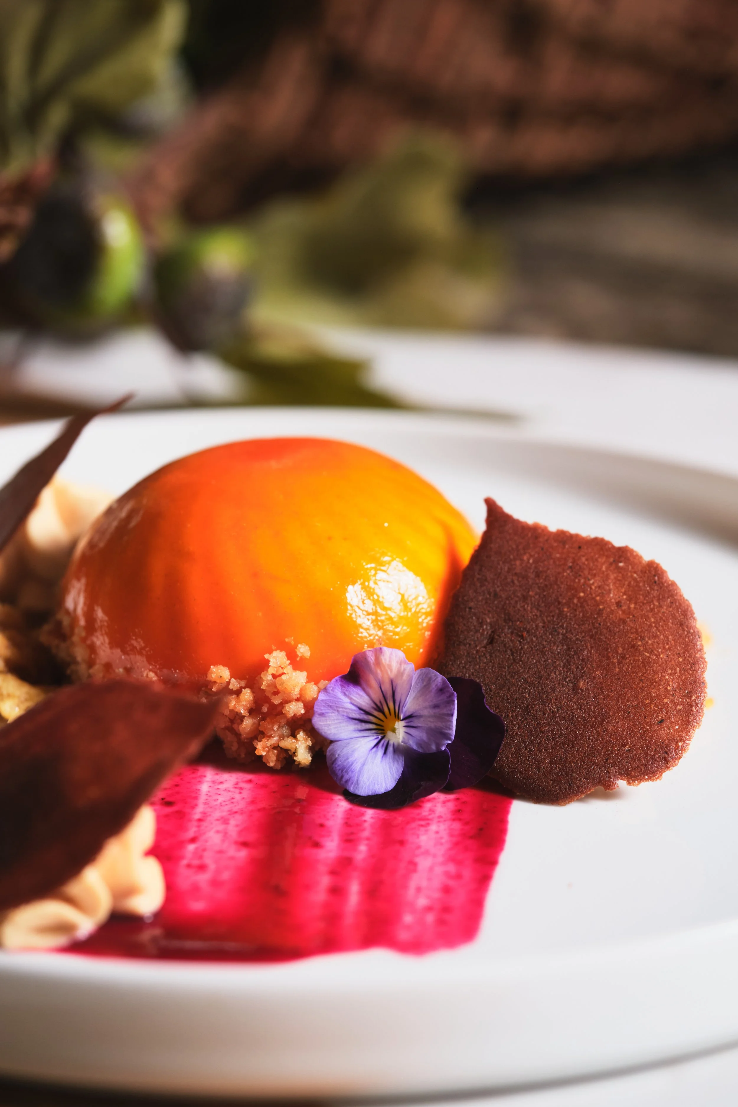 A plated dessert featuring a round, glossy orange and yellow fruit or mousse, garnished with a purple edible flower, chocolate cookie or meringue, and purple and red leaves, with a pink sauce on the white plate.