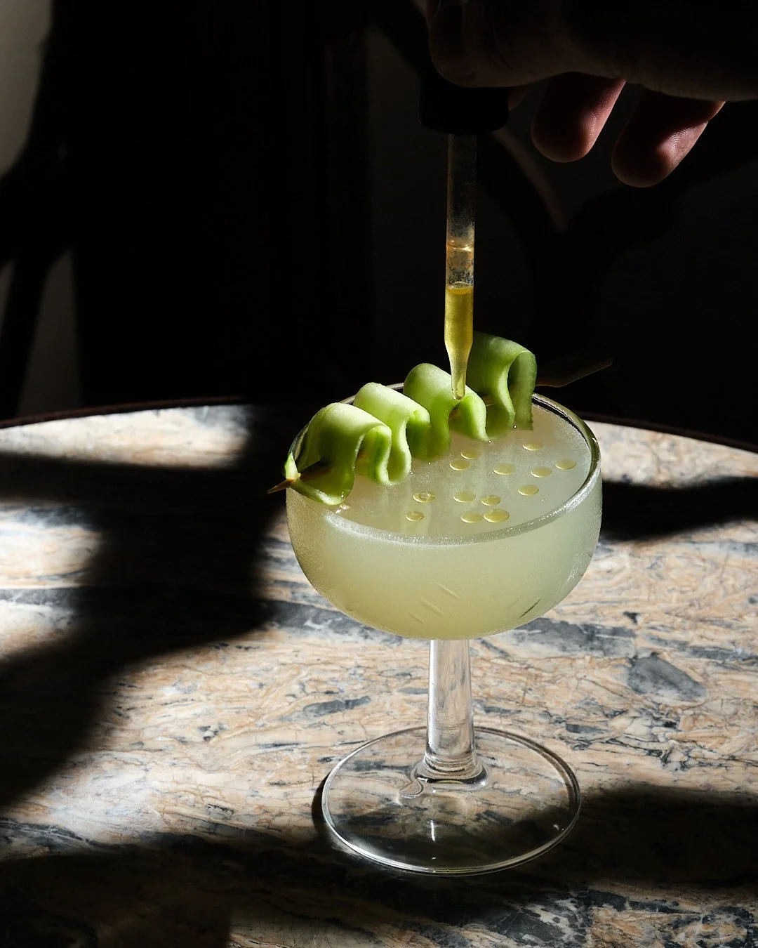 A cocktail glass with a pale yellow-green drink, garnished with rolled green melon slices and a splash of yellow liquid being added with a dropper.