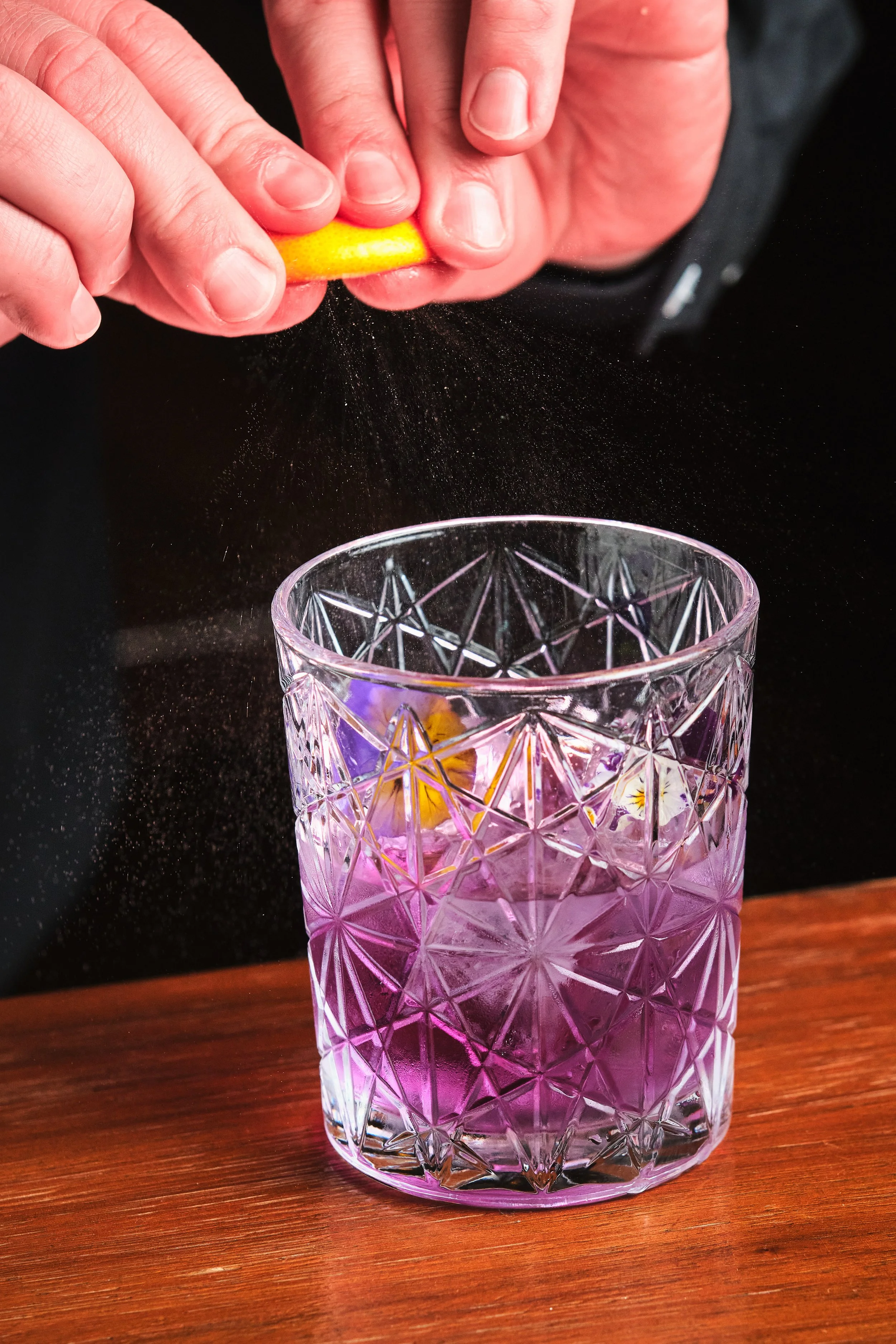 A person grating a lemon over a purple glass filled with ice on a wooden surface.