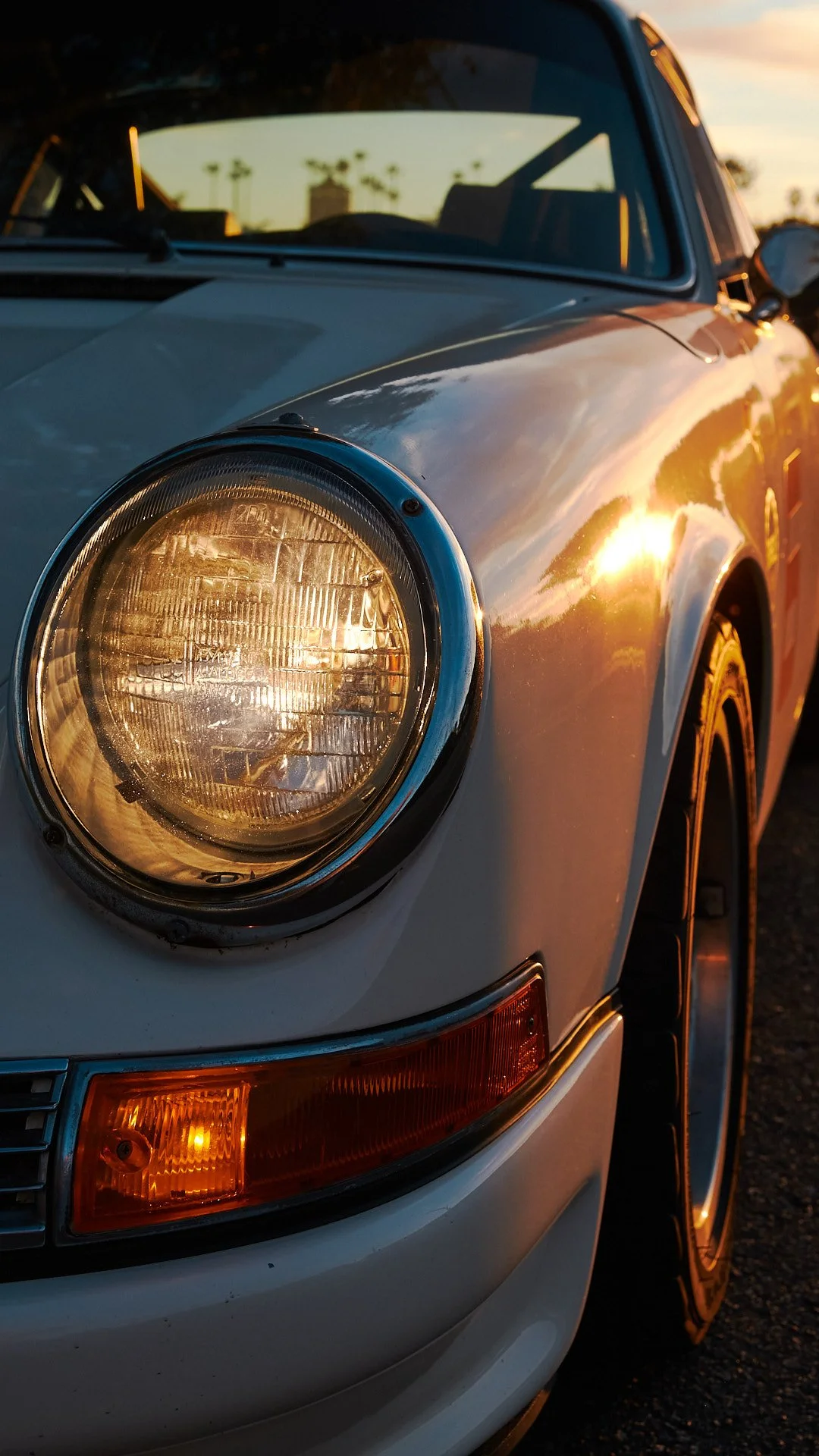 Close-up of a classic white Porsche 911 with the front headlight and turn signal illuminated during sunset.