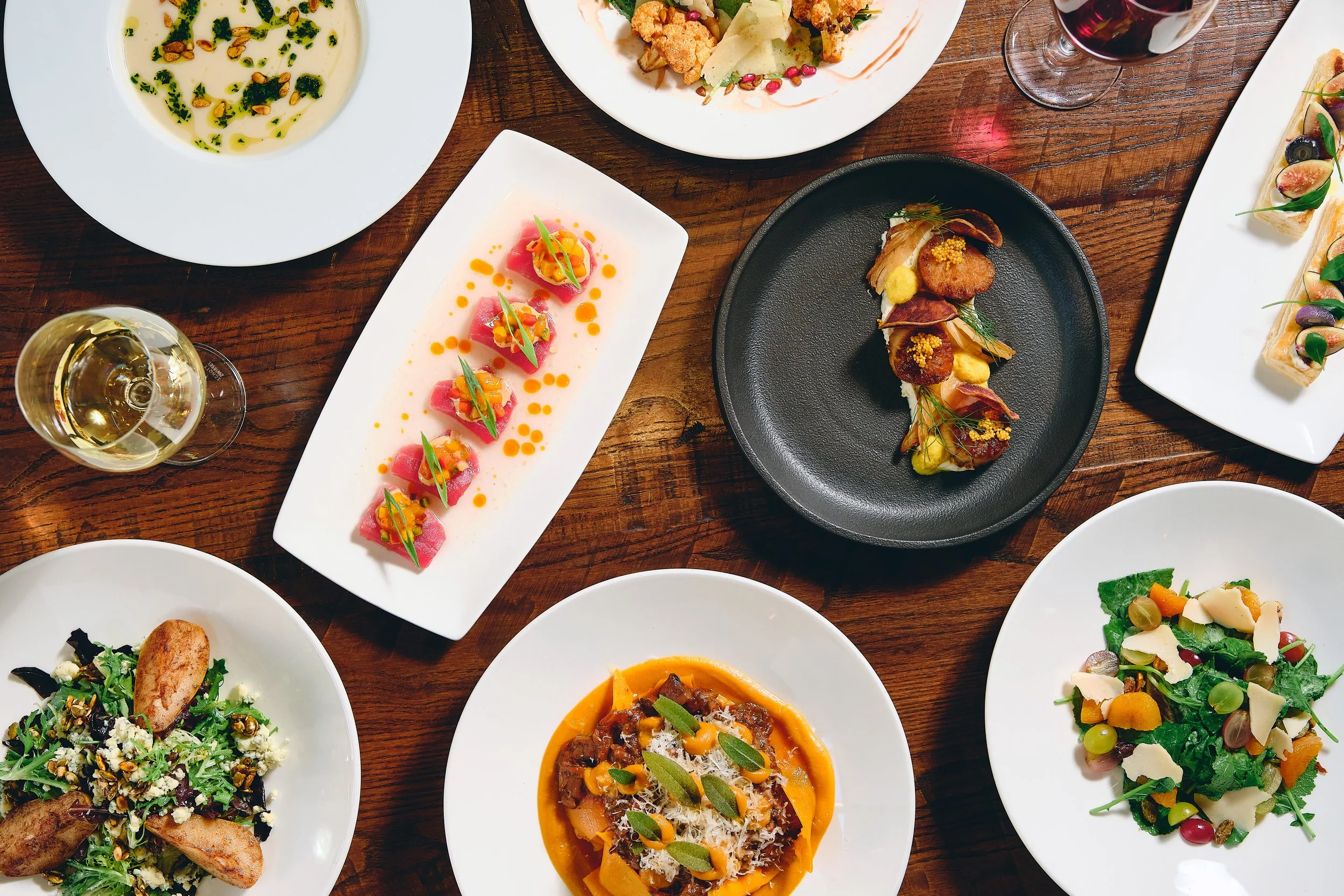 A table with a variety of gourmet dishes, including a plate of sashimi with orange sauce, a fresh salad with potatoes and cheese, and several other plated entrees, along with a glass of white wine and a glass of red wine.