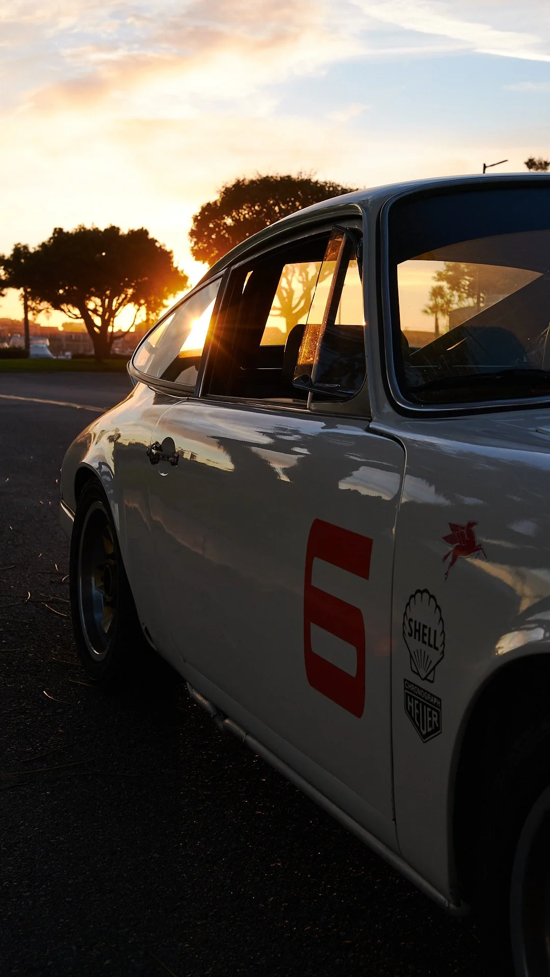 A vintage racing car with the number 6 on its side, parked on a street at sunset, with trees and a partly cloudy sky in the background.