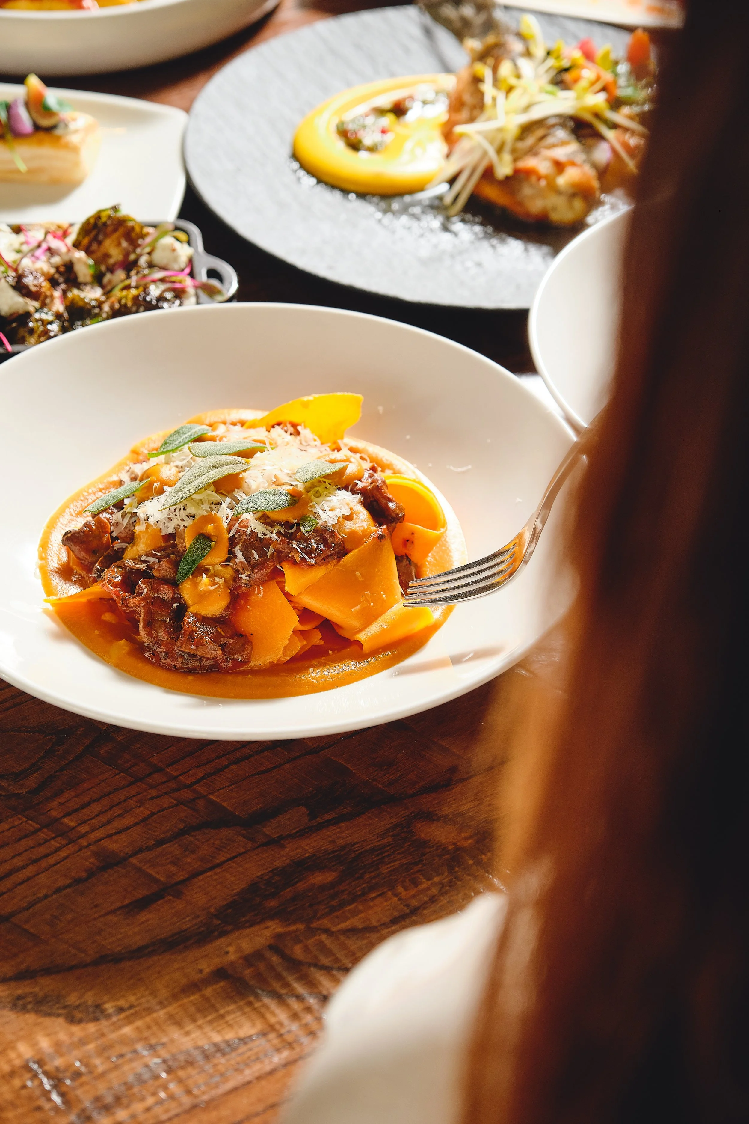 Close-up of a bowl of pasta with orange sauce, garnished with herbs and cheese, on a wooden table with various other dishes in the background.