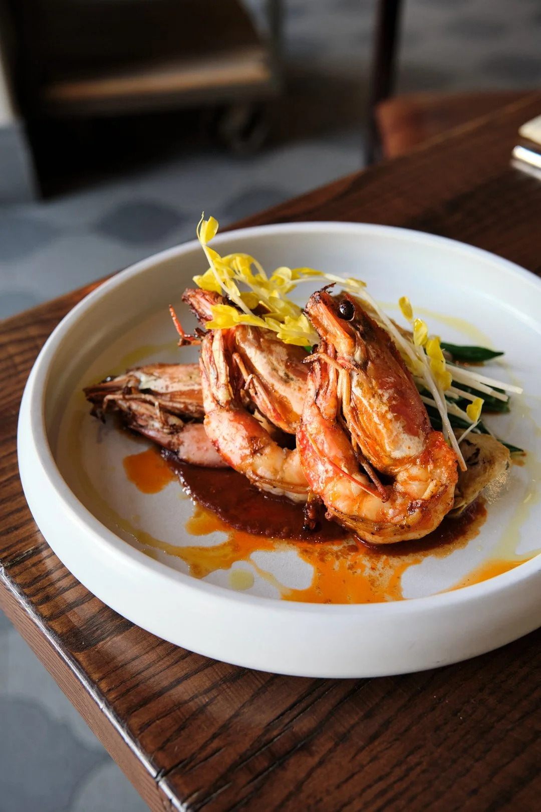 A dish featuring cooked shrimp with sauce, garnished with yellow and green vegetables on a white plate placed on a wooden table.