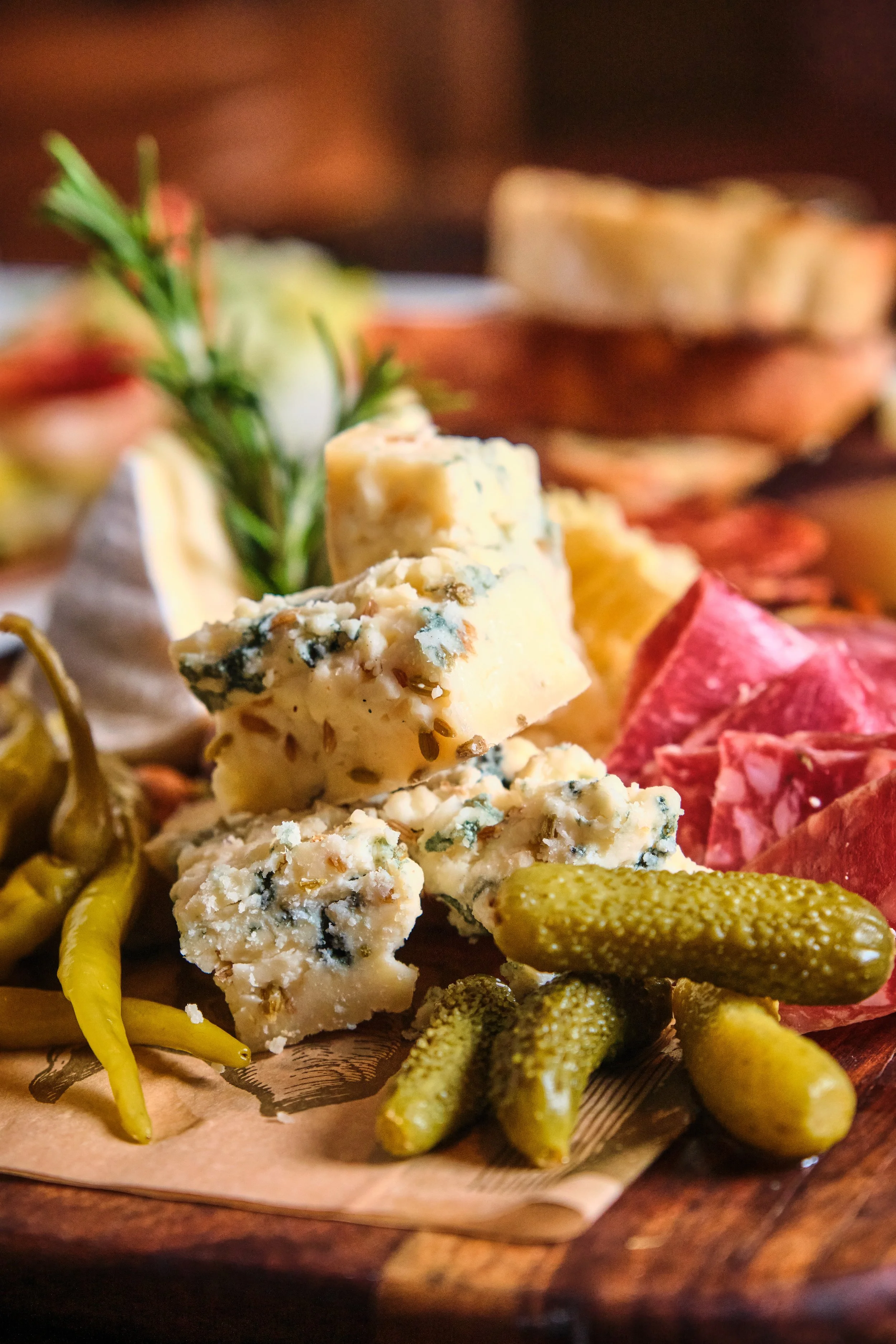 Assorted cheeses, green pickles, and sliced meats on a wooden platter with herbs