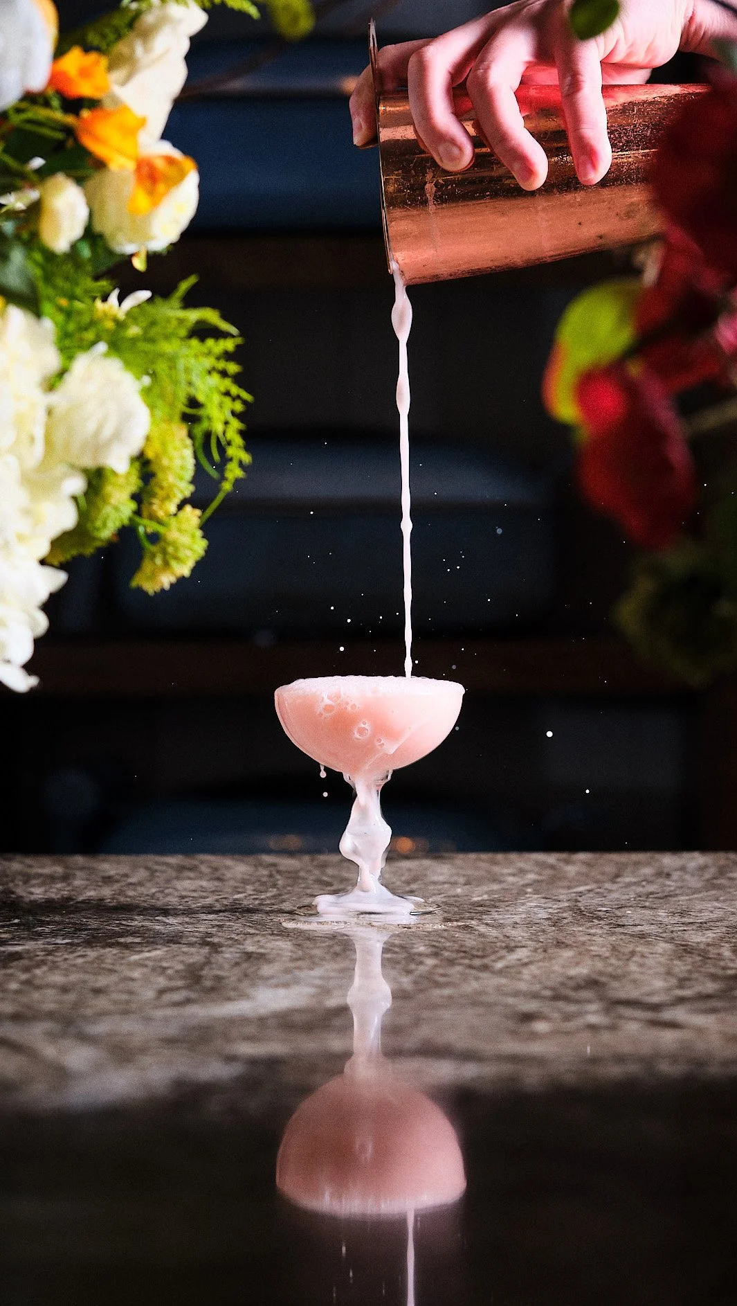 Close-up of a bartender pouring a pink cocktail from a copper shaker into a coupe glass, with flowers on either side and a reflection on a marble surface.