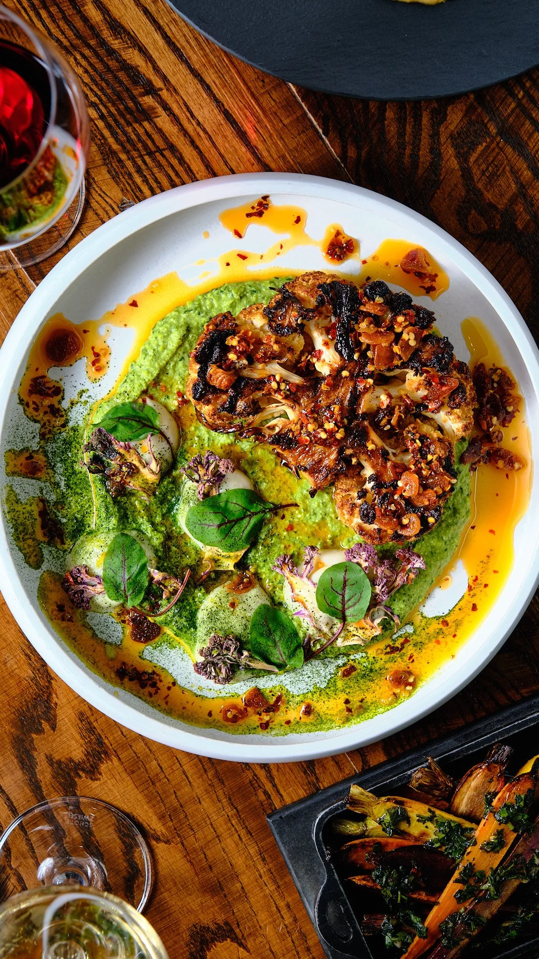 A white plate of roasted cauliflower topped with a spicy chili sauce and garnished with microgreens, resting on a bed of green sauce and surrounded by a yellow oil drizzle, on a wooden table with glasses of wine nearby.