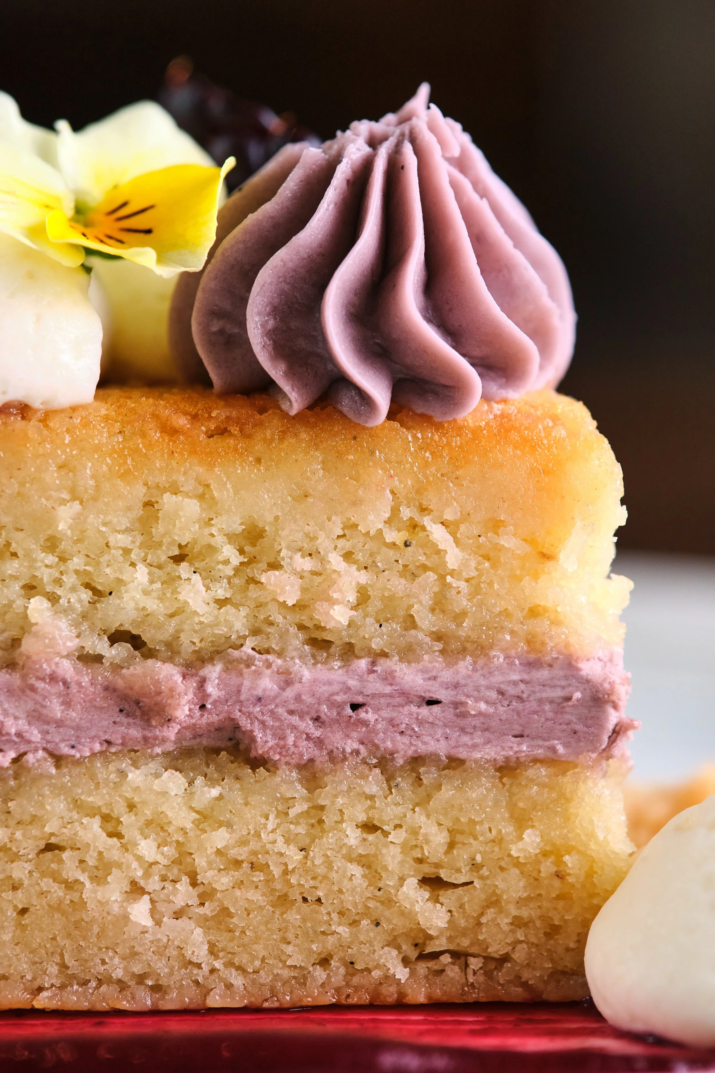 A close-up of a slice of vanilla cake with pink frosting and purple frosting on top, garnished with a yellow flower and dollops of white and purple cream.