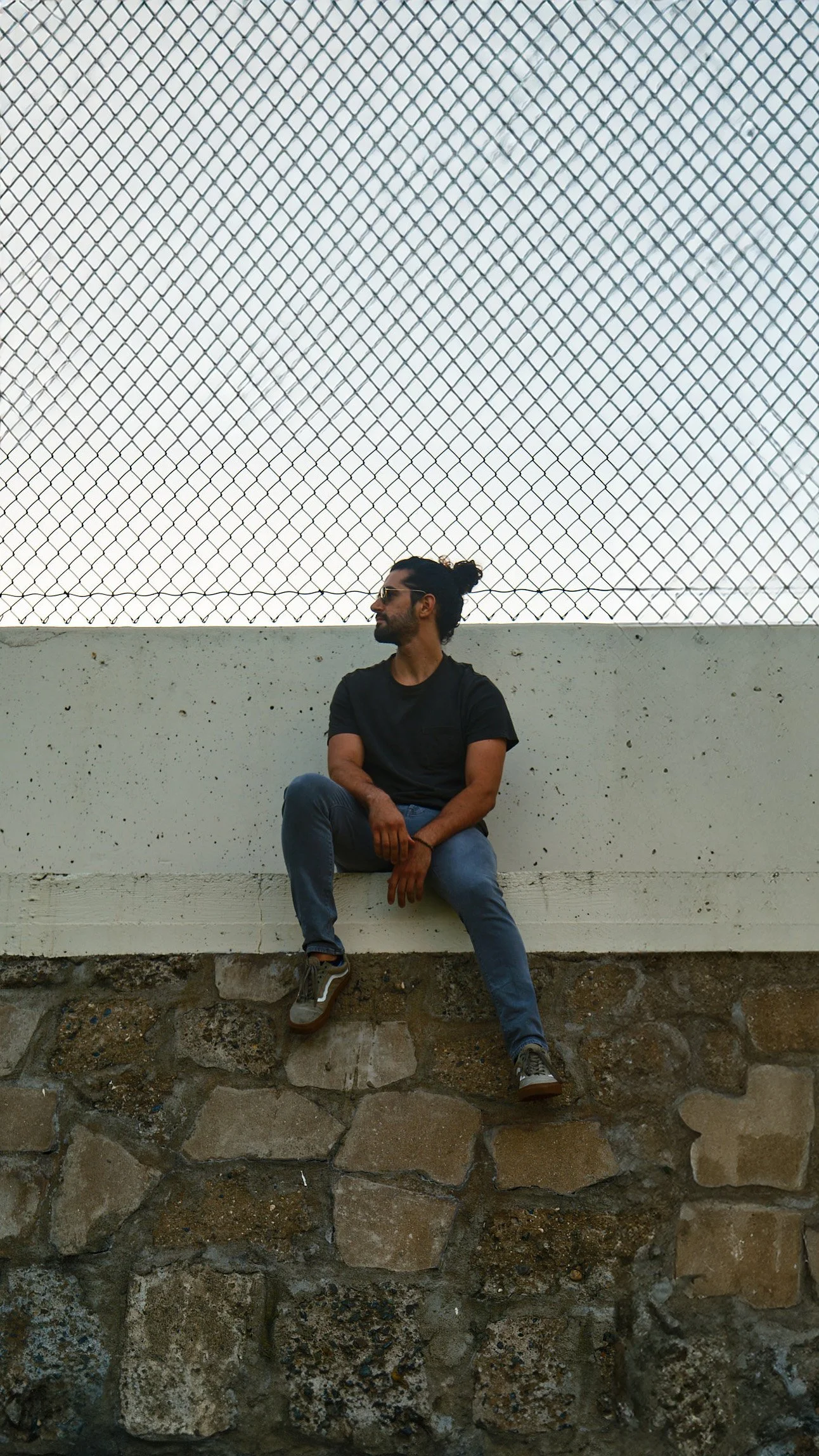 A man in a black T-shirt and blue jeans sits on a low wall with his hands resting on his knee, looking to his left, with a chain-link fence and white wall behind him.