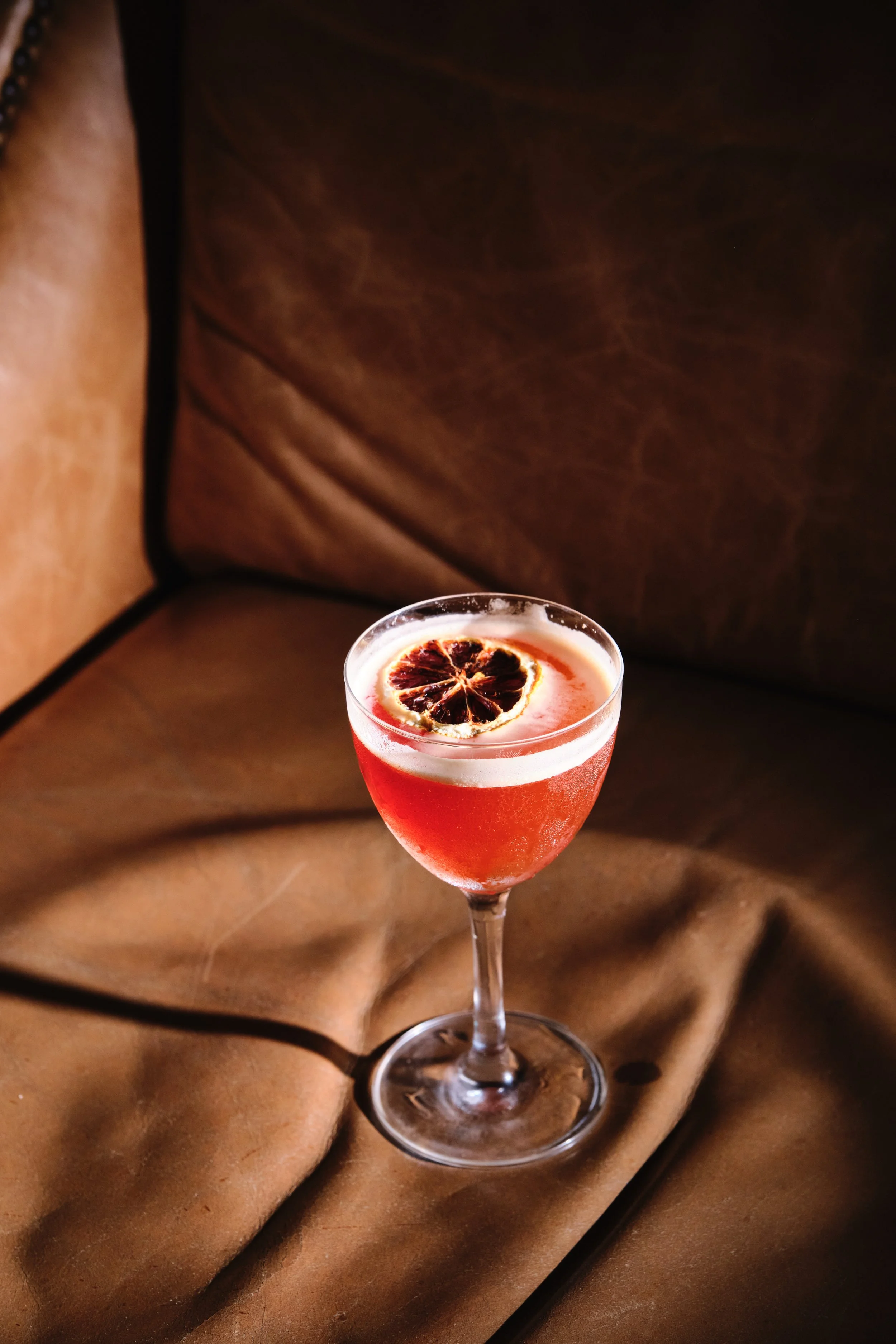 A cocktail in a coupe glass with a dried citrus slice on top, placed on a brown fabric surface.