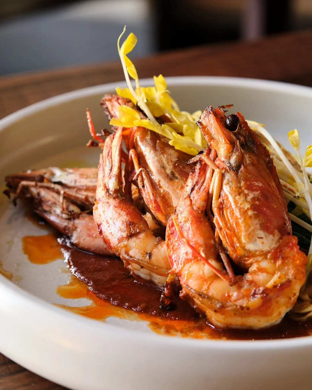 Close-up of grilled shrimp on a white plate with a dark sauce and garnished with yellow shredded vegetables.