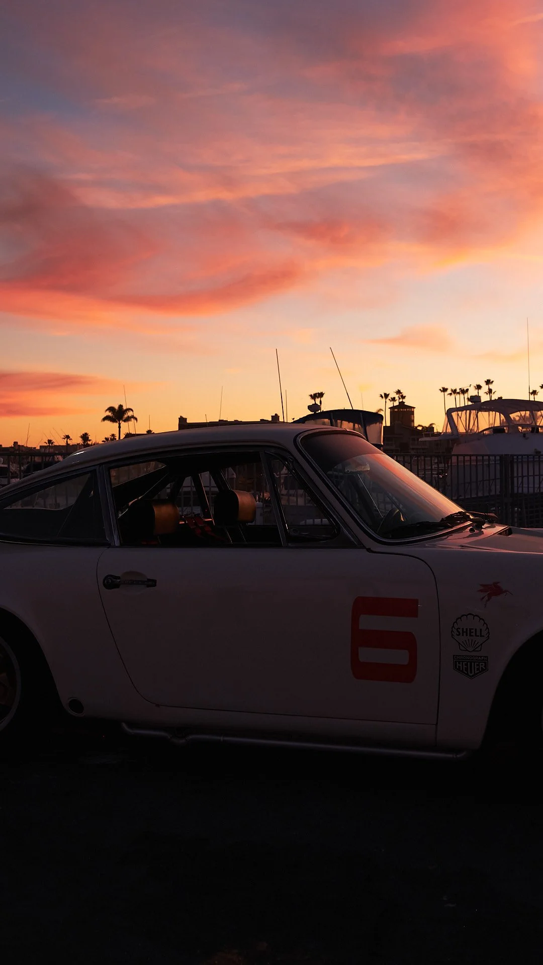 A vintage race car with a large red number 6 on its side, parked near the water, under a colorful sunset sky, with boats and palm trees in the background.