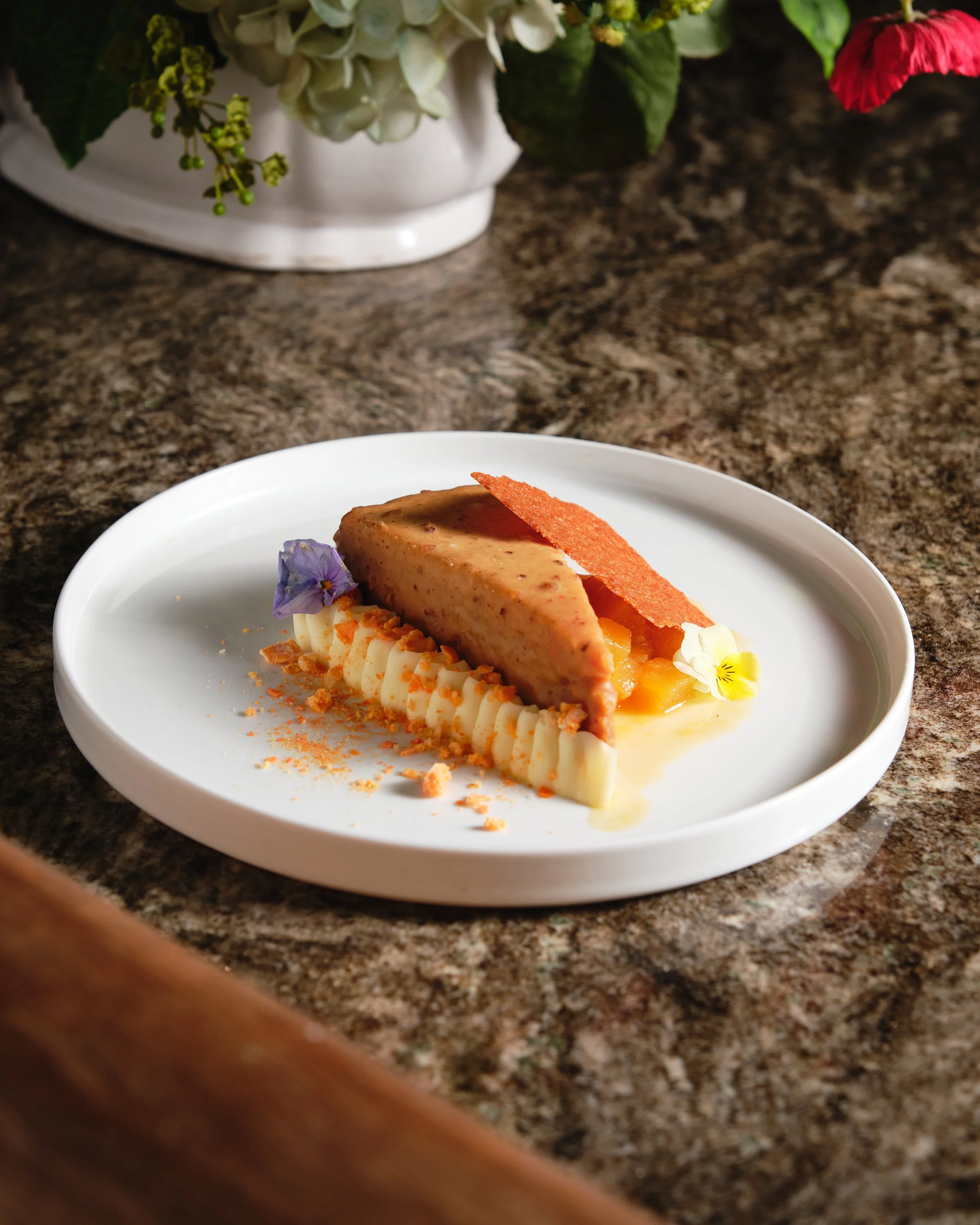 A slice of cheesecake garnished with edible flowers, crumbs, and a caramelized sugar decoration, served on a white plate with a brown speckled countertop and a floral arrangement in the background.