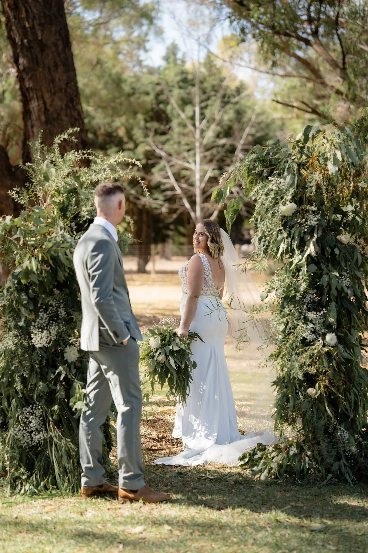 A bride and groom during their outdoor wedding ceremony, standing under a floral arch with green foliage and white flowers. The bride is smiling at the groom, holding a bouquet, and wearing a white lace wedding dress with a train. The groom is dressed in a light gray suit, facing the bride in a wooded area.