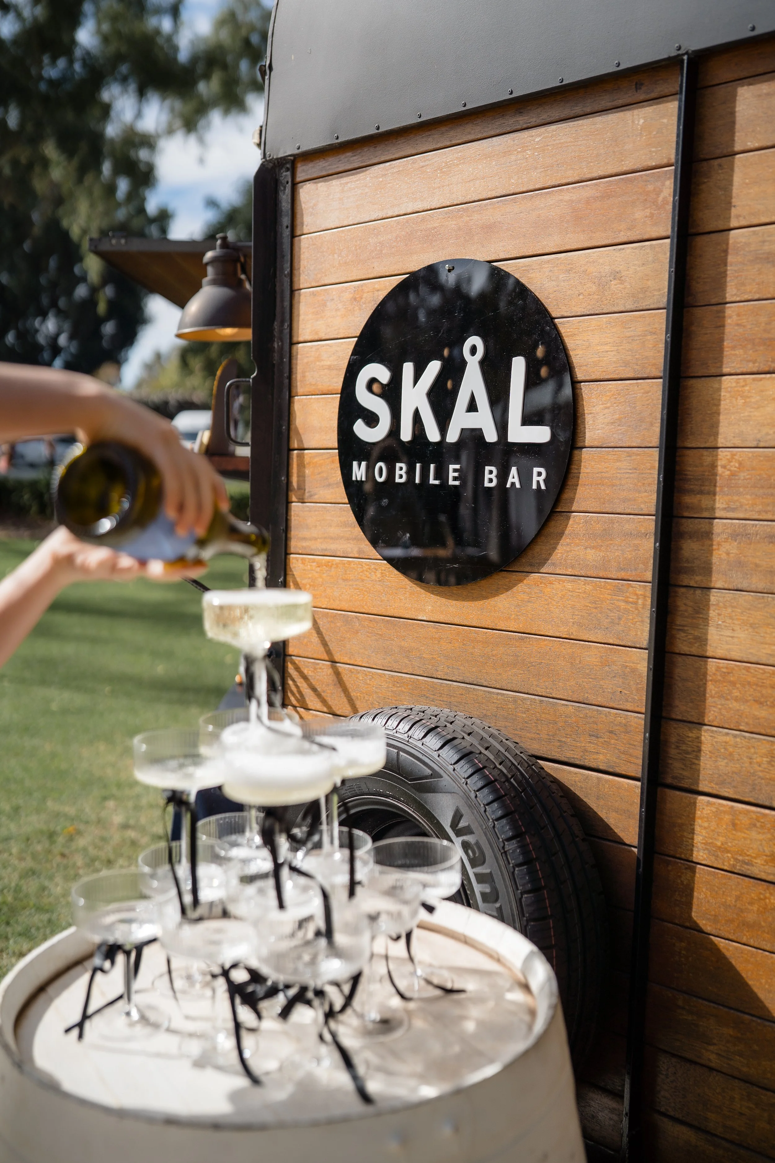 A wooden mobile bar trailer with a round black sign that says 'SKÅL' and 'MOBILE BAR'. In the foreground, there is a multi-tiered drink display with several empty champagne glasses, and someone is pouring a drink into the top glass. In the background, there are trees and a vehicle, suggesting an outdoor setting.