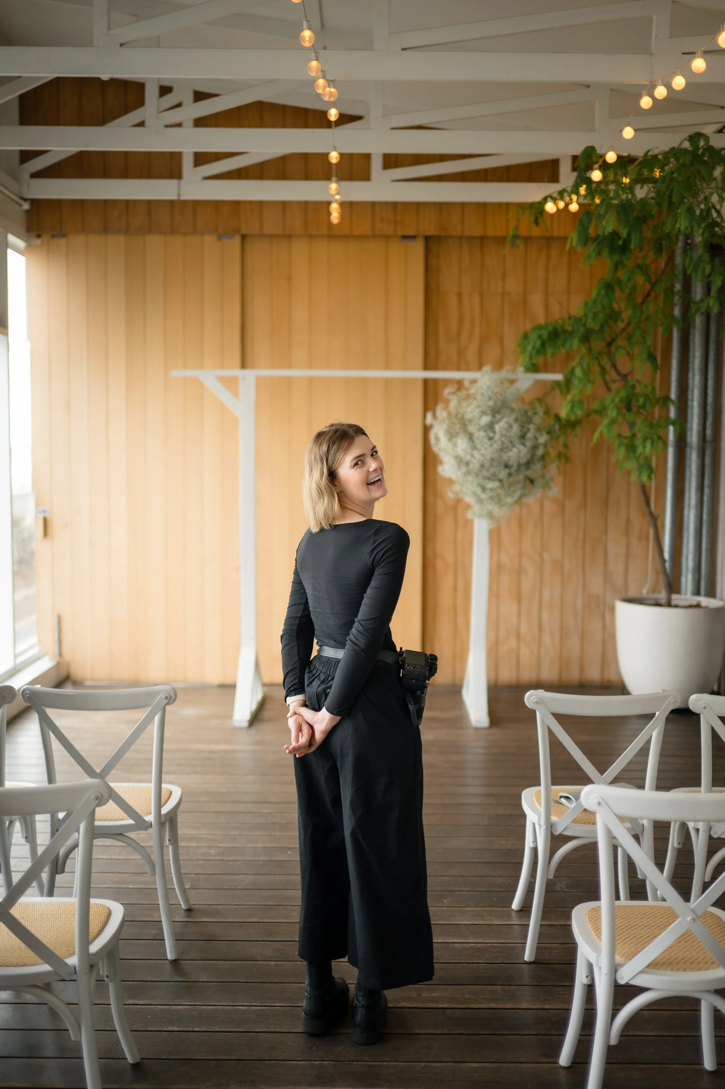 A woman wearing a black long-sleeved top and wide-leg black pants standing indoors in a decorated space with string lights, white chairs, and potted plants, smiling and looking over her shoulder.