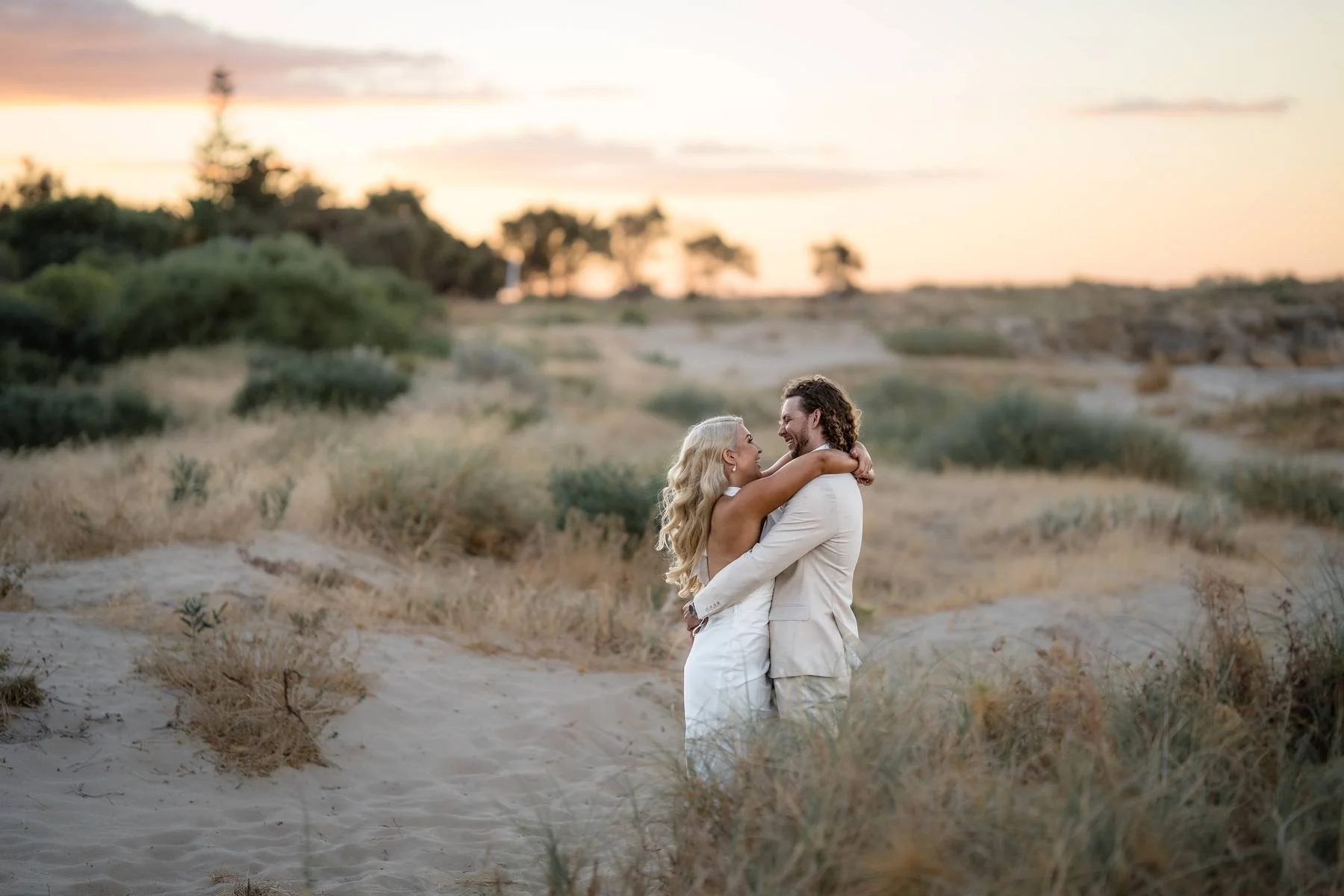 A couple embracing and smiling at each other in a sandy, grassy outdoor setting during sunset.