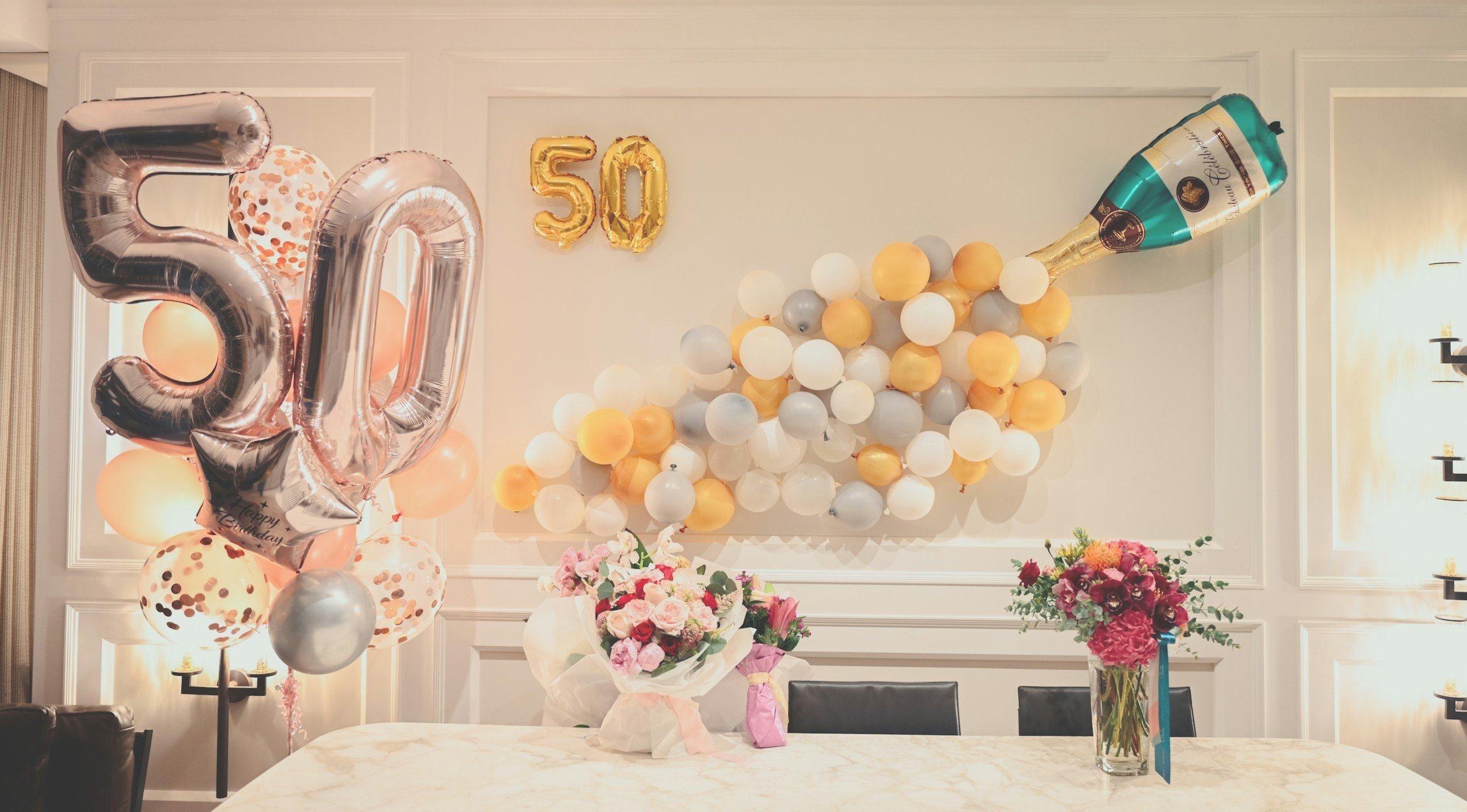 Decorative 50th birthday celebration setup with large metallic balloons reading '50', a cluster of pink, white, yellow, and gray balloons, a champagne bottle-shaped balloon, and two flower bouquets on a white table in a well-lit room.
