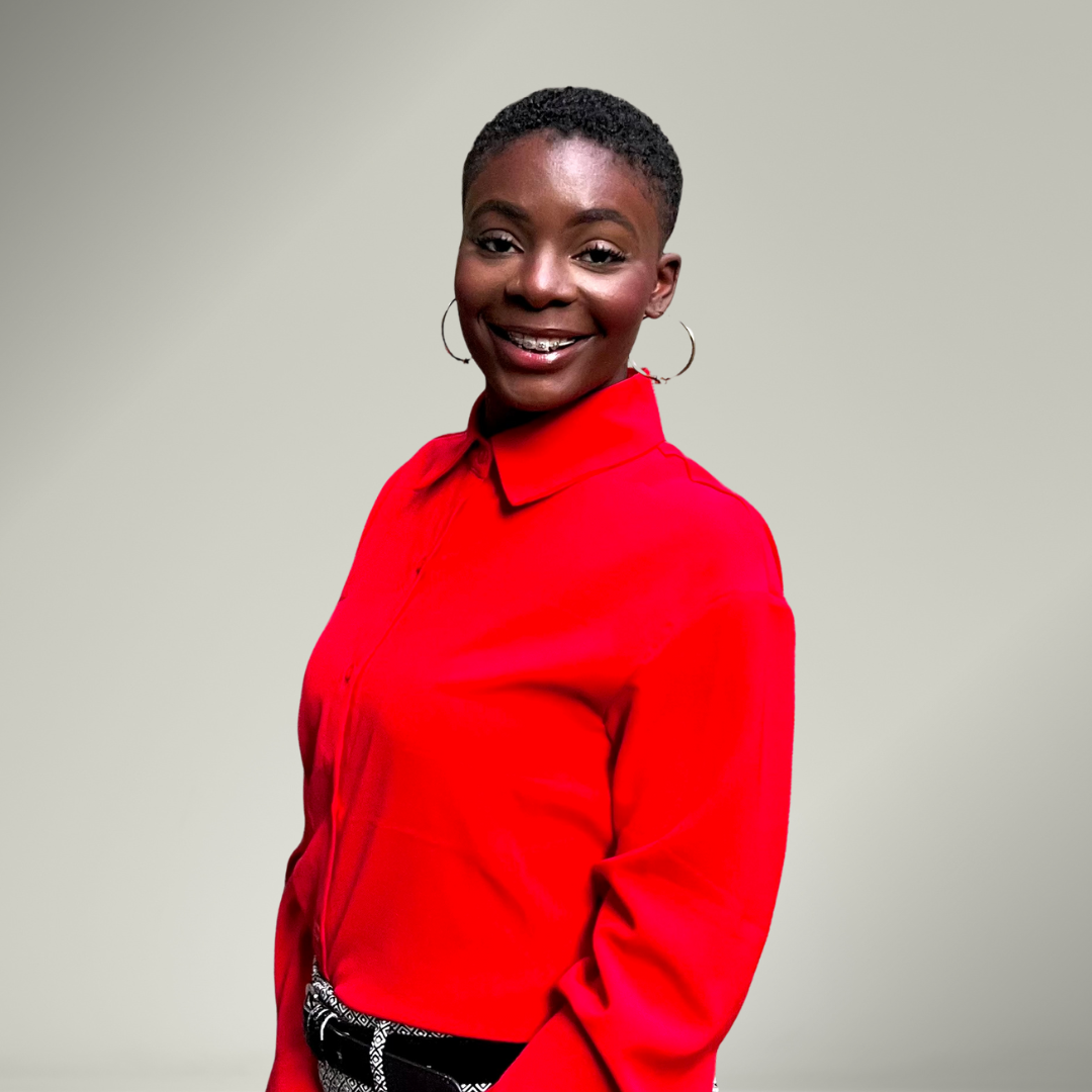 A smiling Black woman with short hair wearing a bright red button-up shirt and hoop earrings, standing against a plain light-colored background.