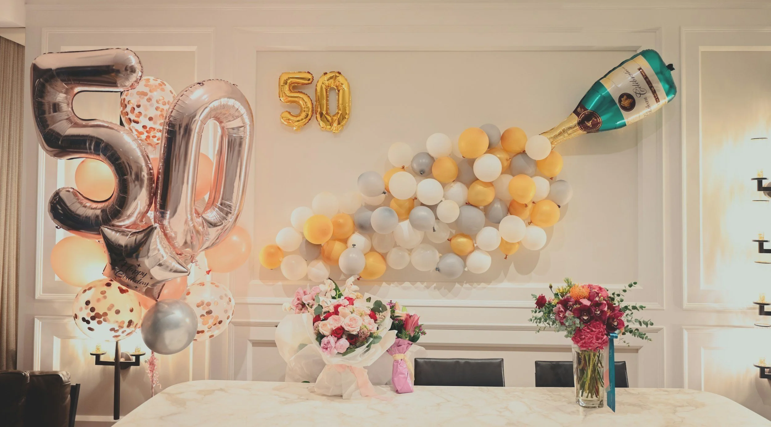 A celebration setup with large silver and pink balloons creating the number 50, a cluster of balloons in white, gold, gray, and peach, a large champagne bottle balloon, and floral arrangements on a table.