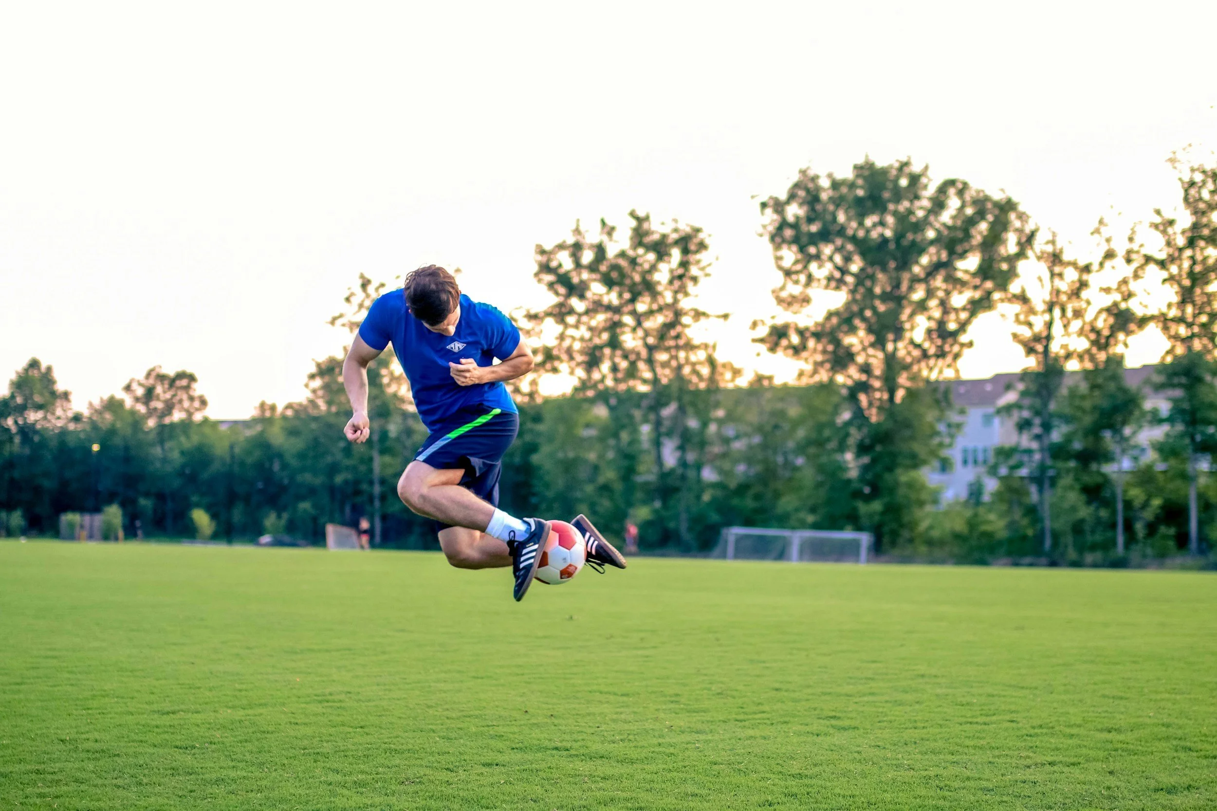 A young man in a blue shirt and shorts playing soccer on a green field during sunset.
