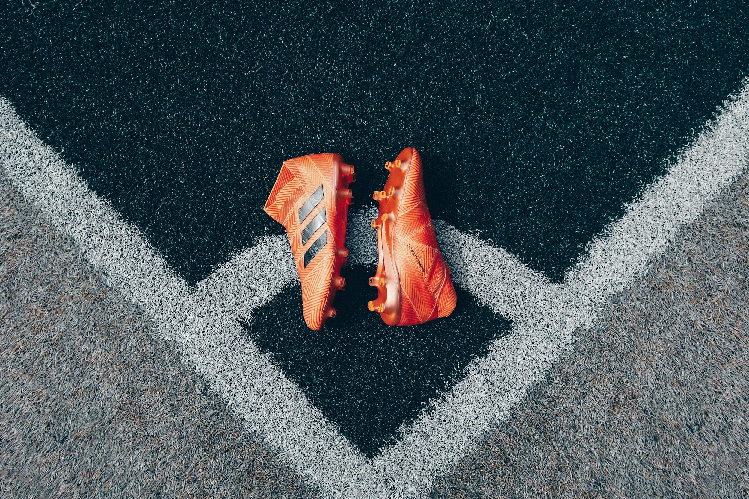 Orange and red Adidas soccer cleats placed on a black and gray turf field at a corner of the goal line.