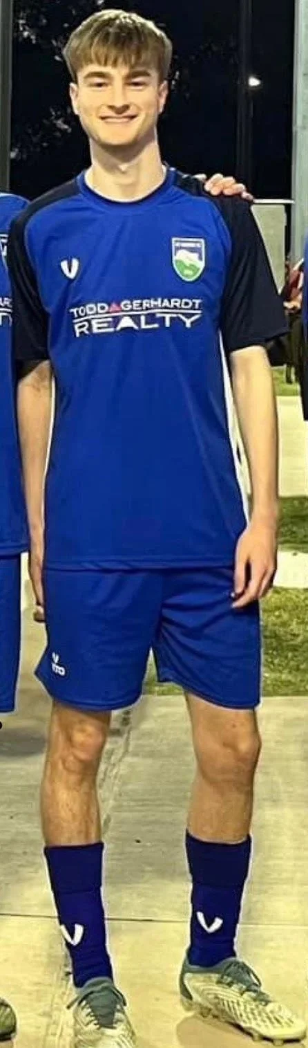 Young man in blue soccer uniform standing outdoors at night, smiling, with another person’s hand on his shoulder.