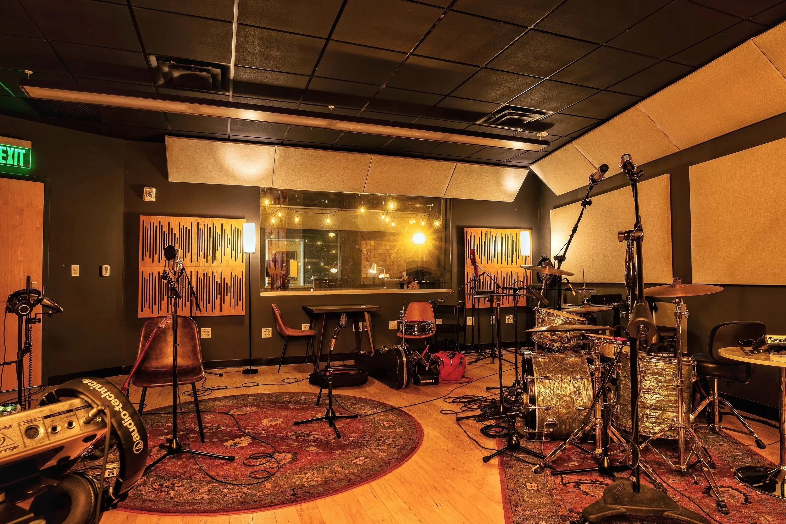 Music recording studio with drums, microphones, chairs, and soundproofing panels, illuminated with warm lighting.