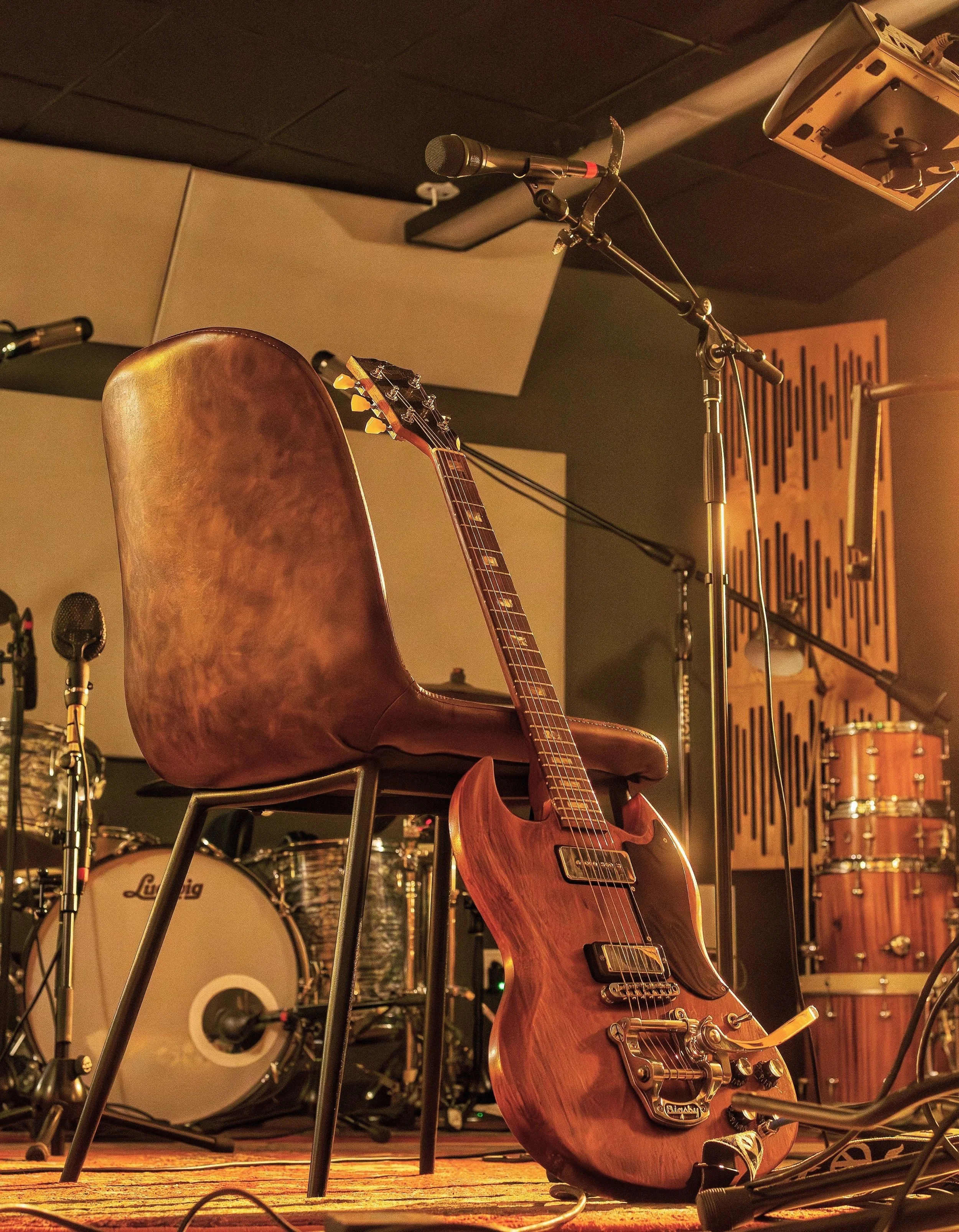 A stage setup with a wooden electric guitar resting on a chair, a microphone on a stand, and a drum set in the background.