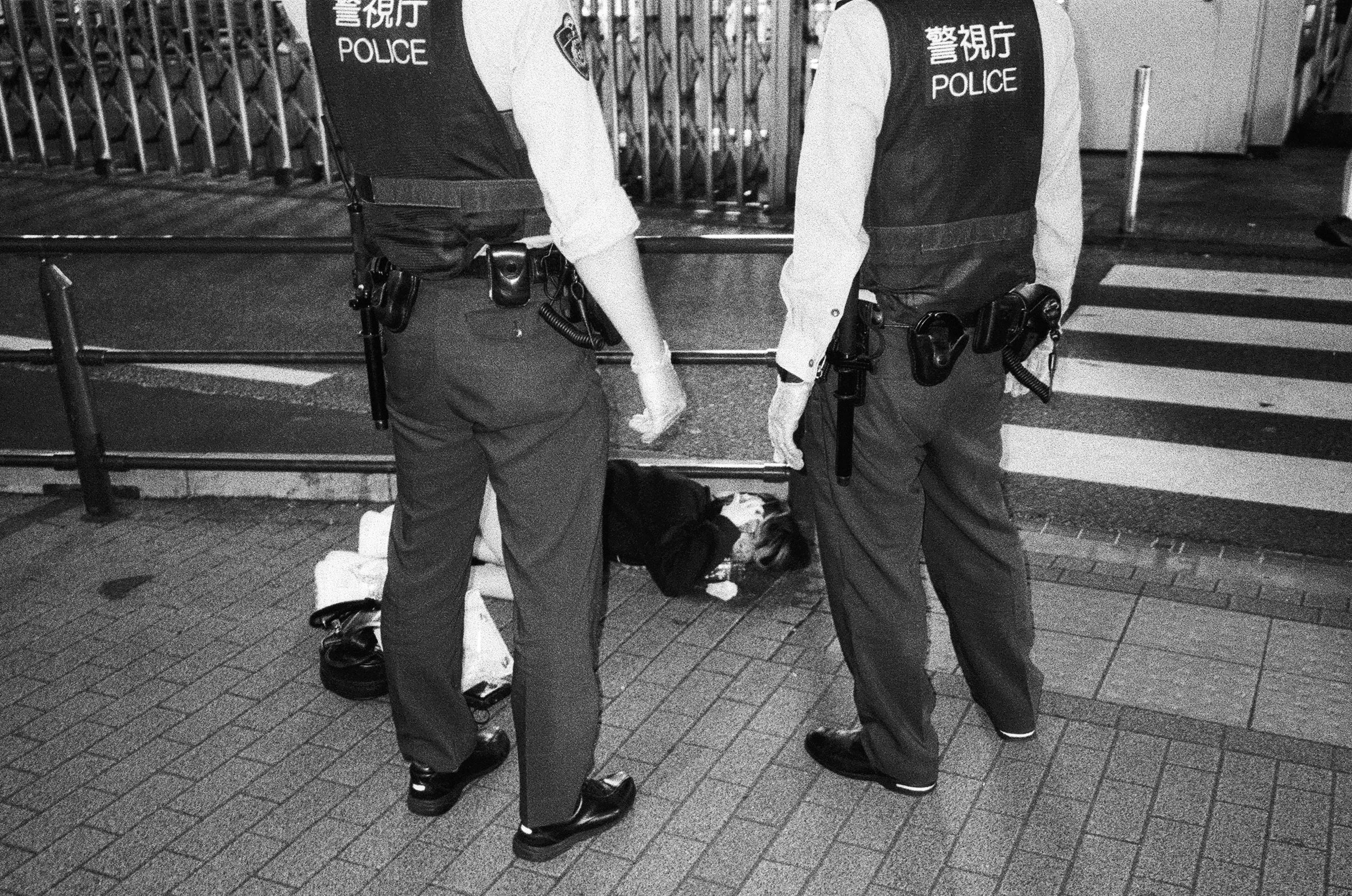 Two police officers are standing next to a person lying on the ground in front of a metal barricade. The officers are facing away from the camera, wearing uniforms with the word 'POLICE' on their backs, and the person on the ground appears to be unre