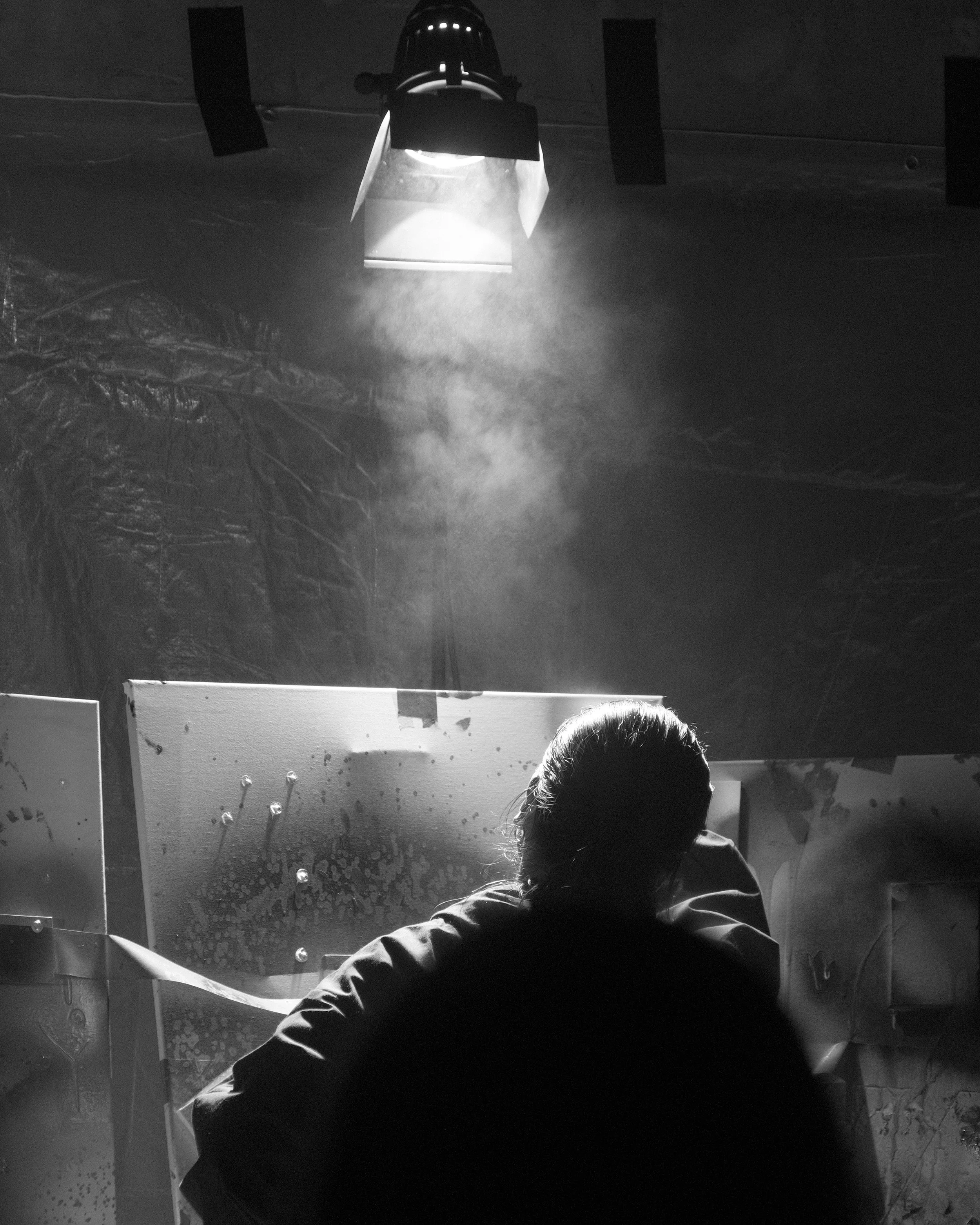 A person working on a painting or art project in a dimly lit room with a bright overhead light and a cloud of dust or spray in the air.